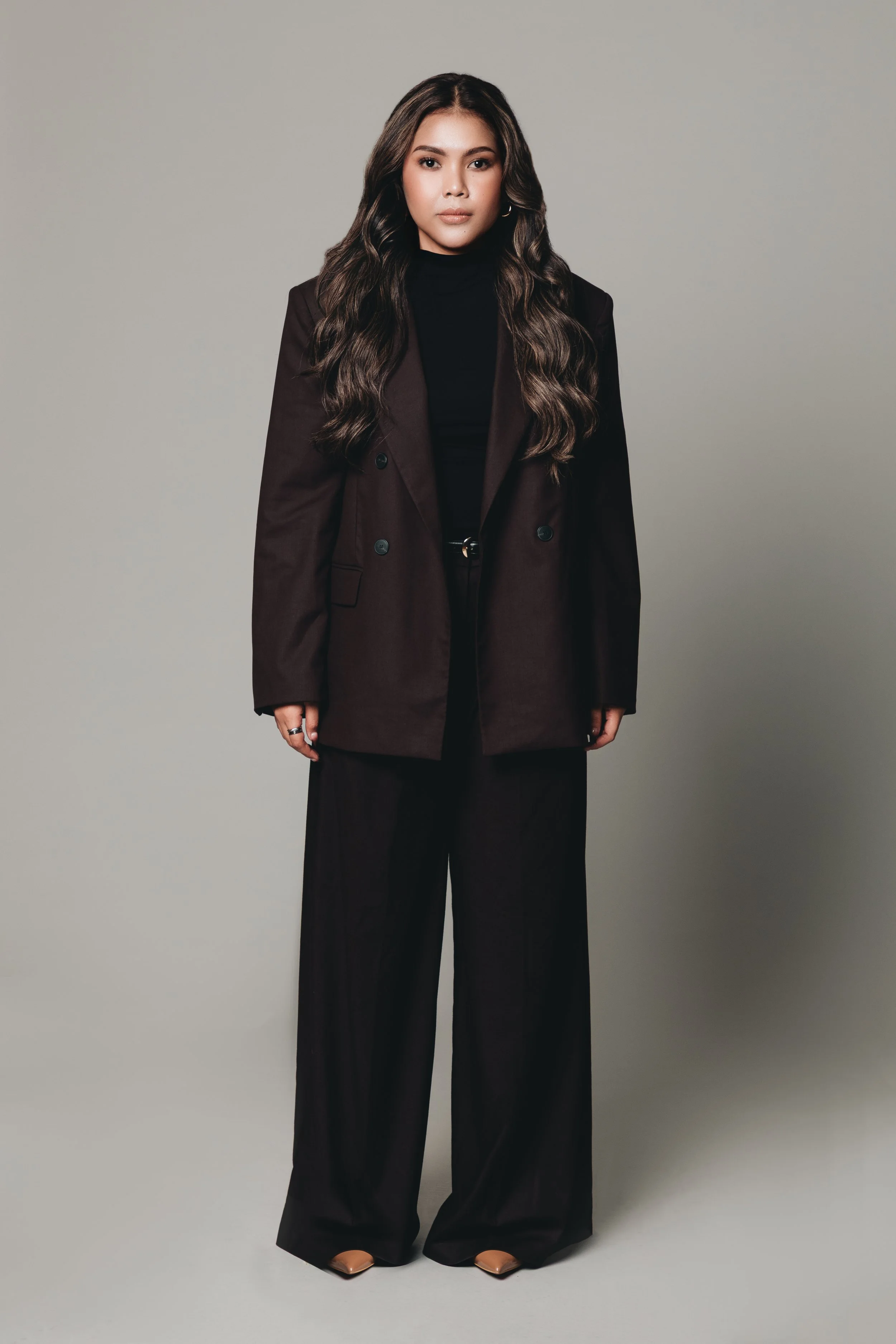 A woman with long wavy hair wearing a black turtleneck, dark brown blazer, and black wide-leg pants, standing against a plain light grey background.