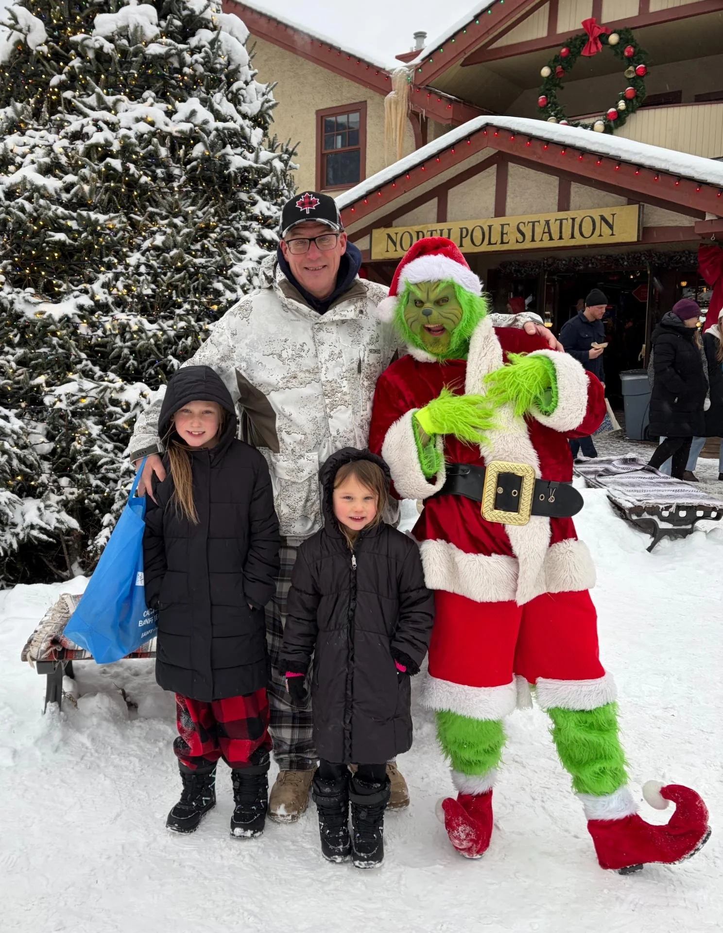 Randy stands with his two granddaughters beside a person dressed as the Grinch in a snowy holiday setting.