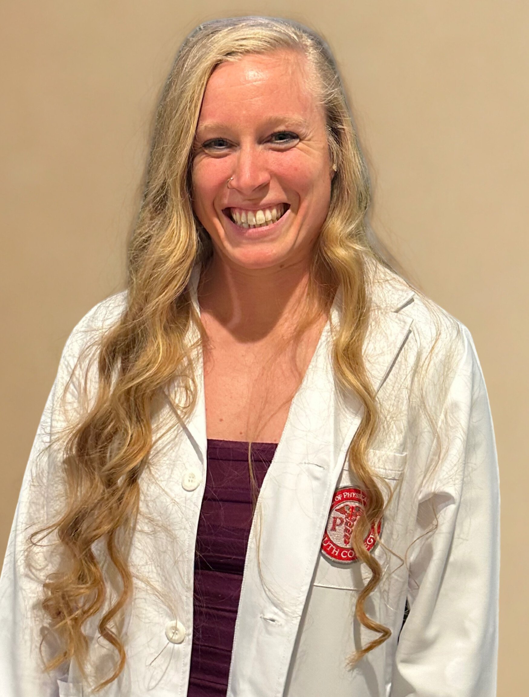 A woman with long, curly blonde hair and a nose ring smiling, wearing a white lab coat with a red emblem on the chest, standing against a beige background.