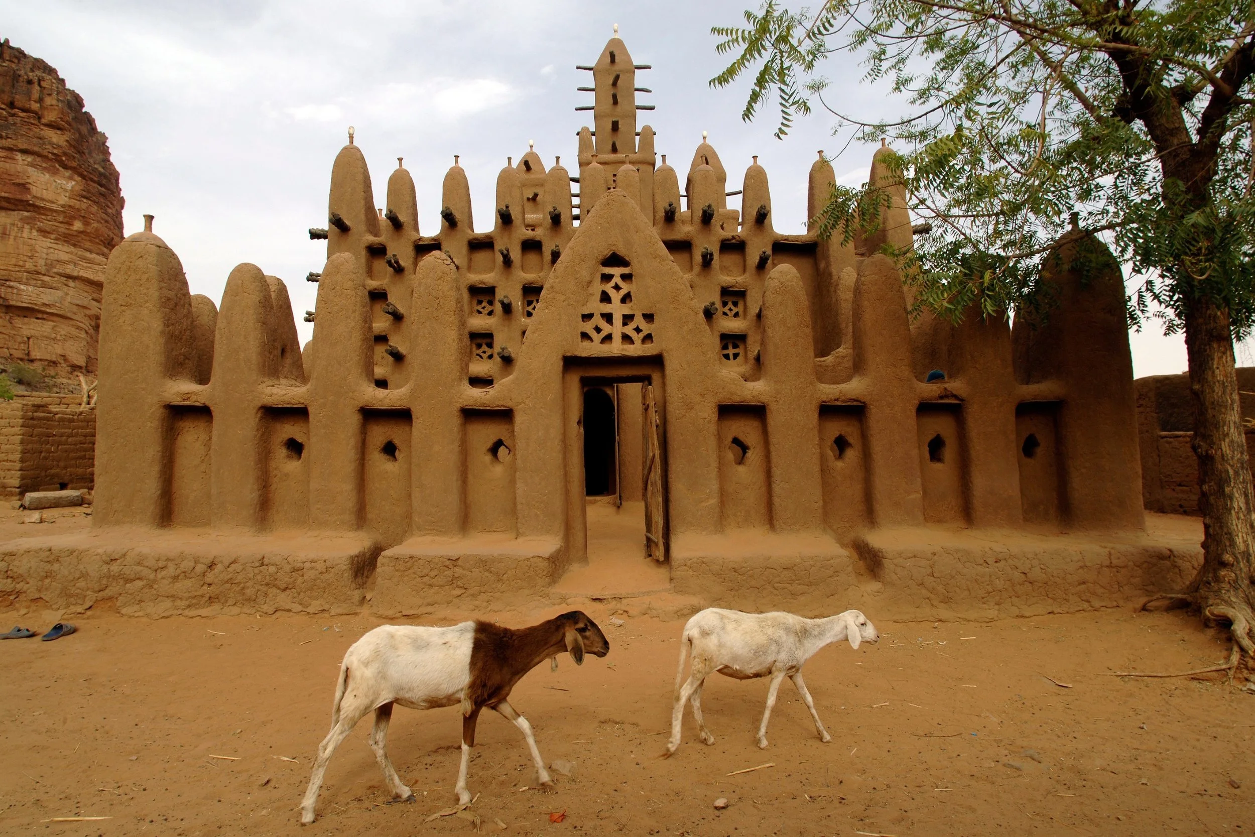 Une mosquée en adobe sur fond de ciel nuageux, avec deux chèvres qui marchent devant, un arbre à droite