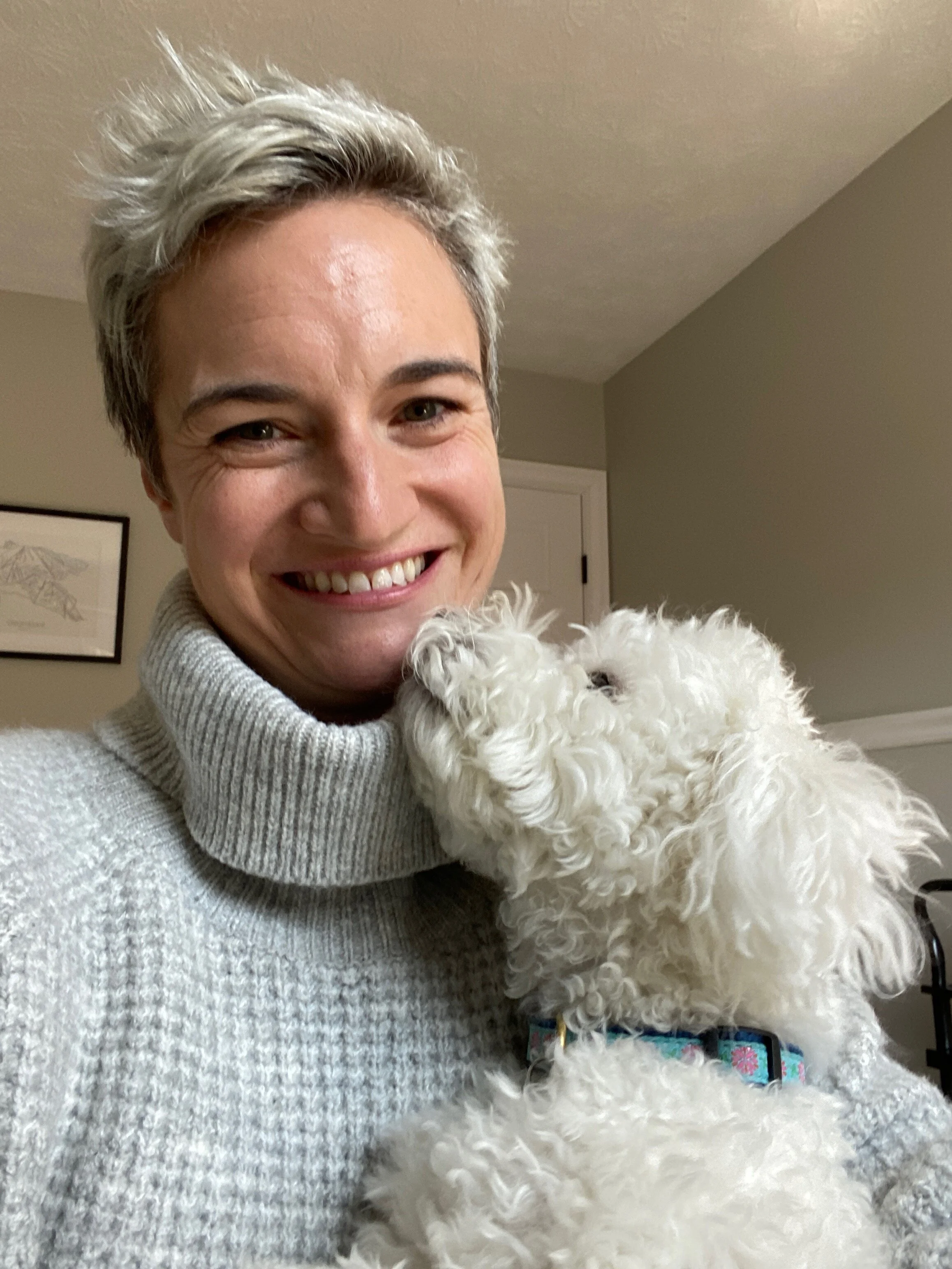 A friendly, happy and nice professional organizer who works with clients, smiling while holding a small, fluffy white dog that is licking their face in a cozy room.