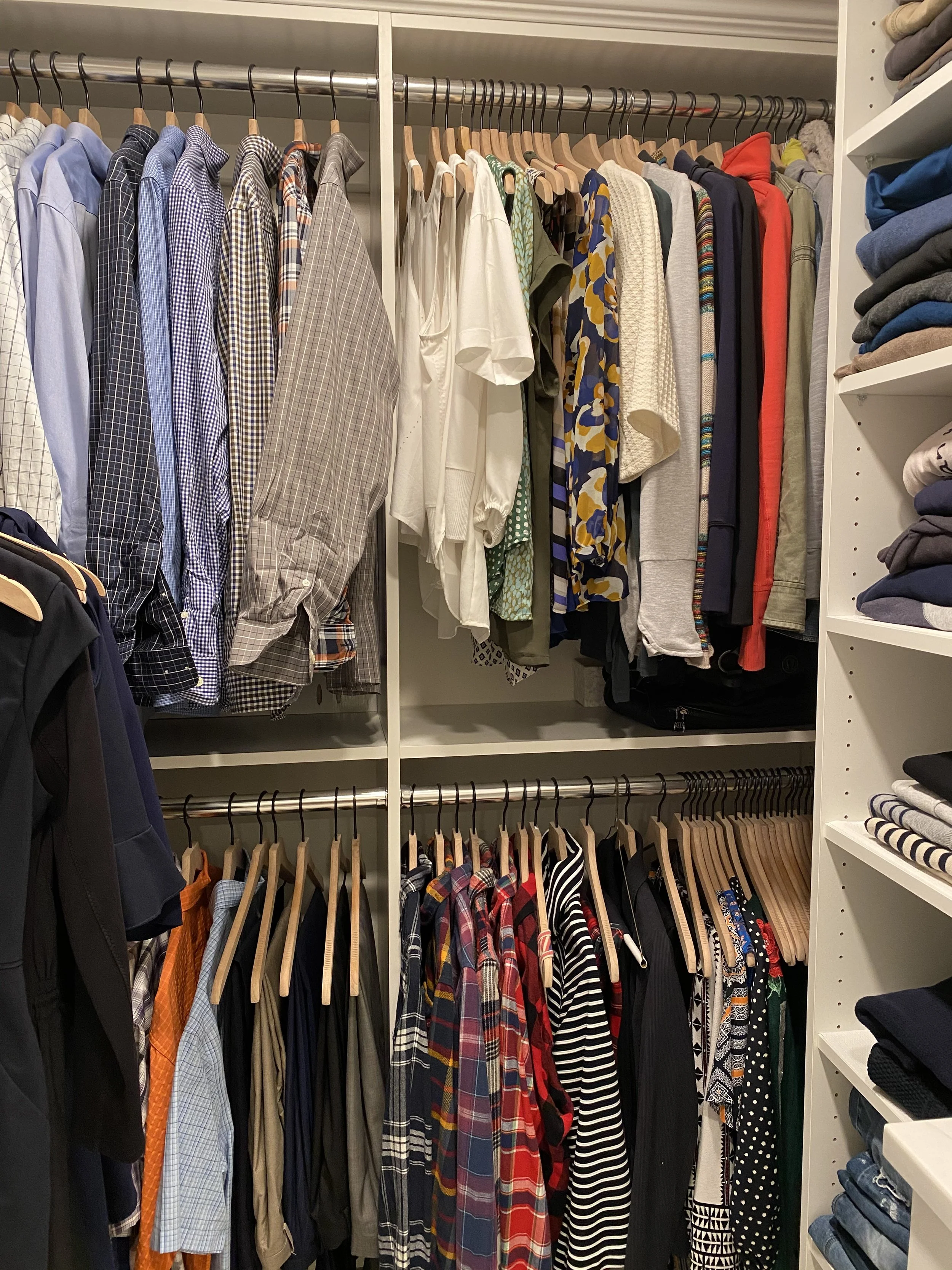 An organized closet with neatly hung shirts, pants, and jackets on wooden hangers, arranged on two horizontal rods inside a white shelving unit, with folded clothes on shelves to the side.