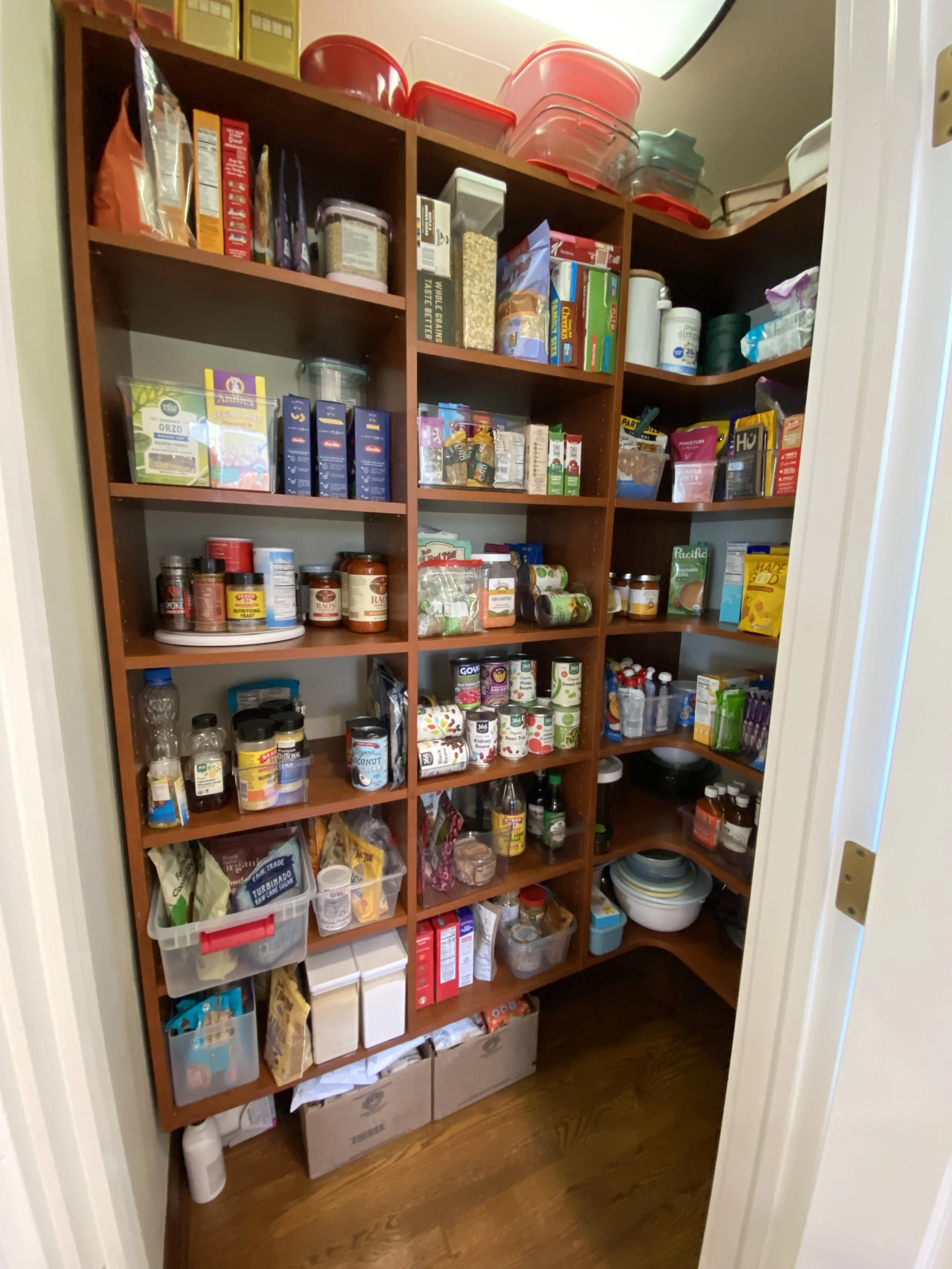 An organized pantry filled with various canned goods, snacks, spices, and kitchen supplies in a small storage closet, using storage bins, containers and shelving systems