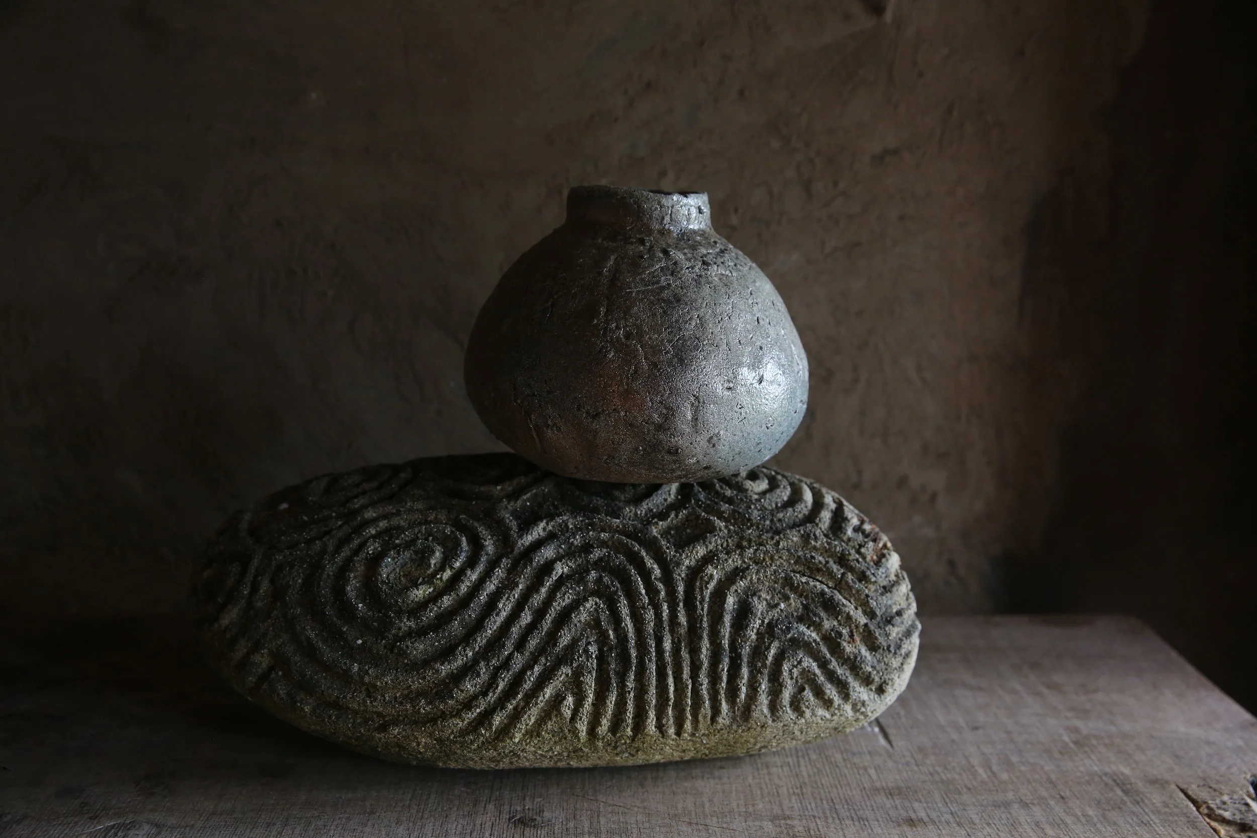 A round, rustic clay pot perched on a textured, segmented stone with an ancient fingerprint pattern, set against a dark, earthy background.