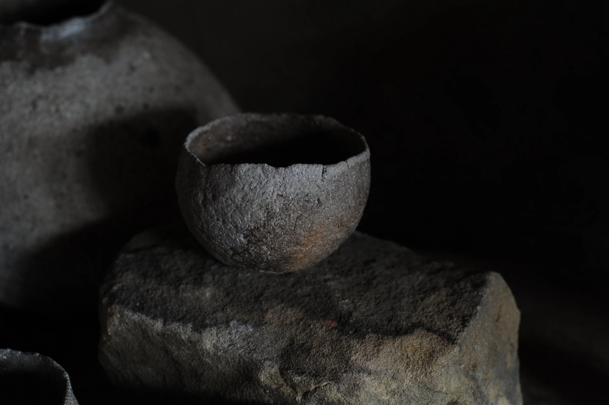 A small, rough-textured bowl made of stone or clay sits on a larger, weathered stone surface against a dark background.