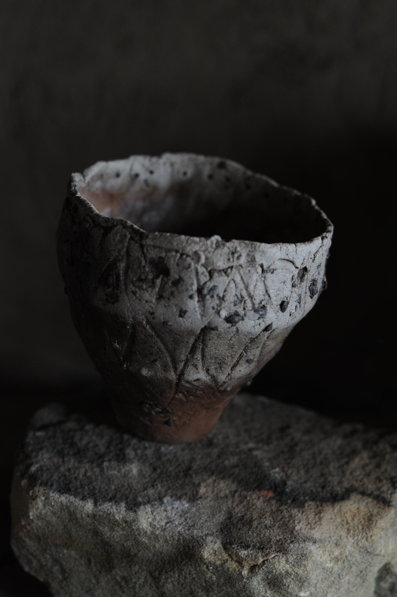 A small, ancient clay cup with carvings on the surface, sitting on a rough stone surface against a dark background.