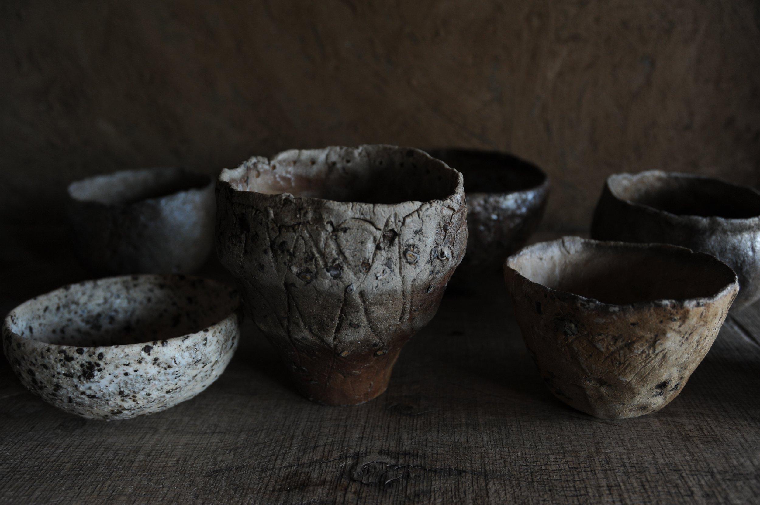 Six rustic, handmade ceramic bowls of various earthy tones and textures arranged on a wooden surface with a dark background.