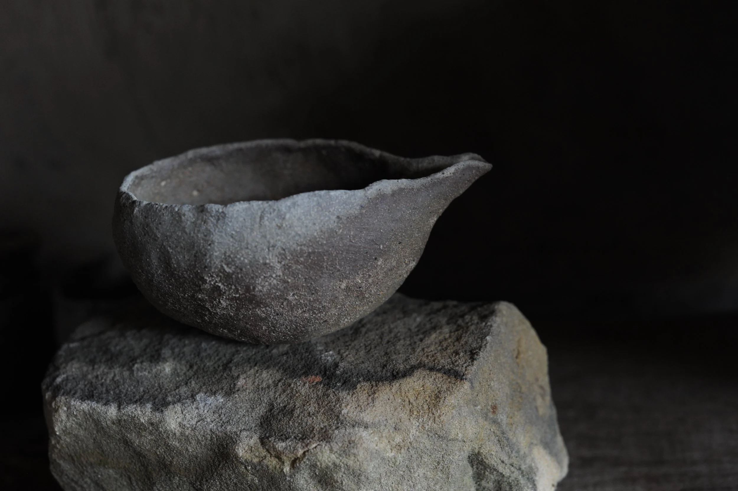 A small, rough textured, gray stone bowl resting on a larger, irregularly shaped stone against a dark background.