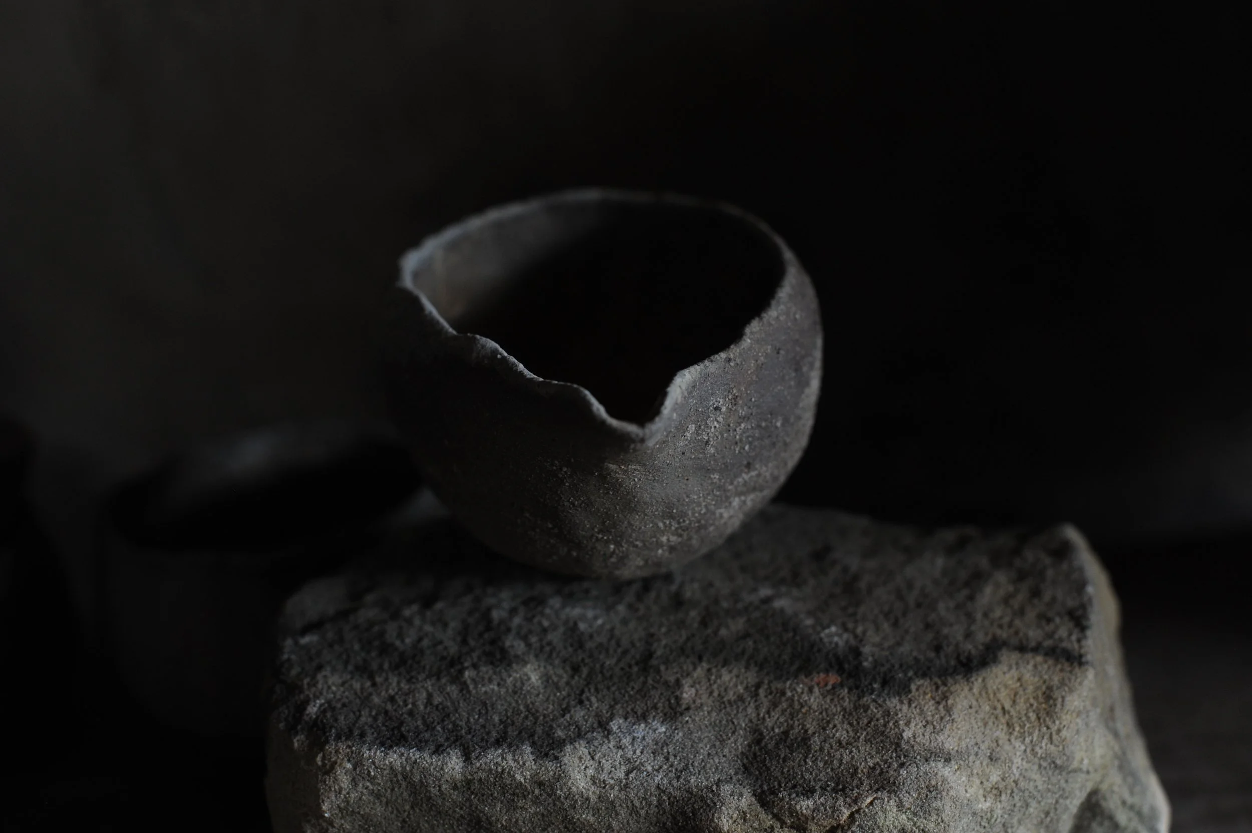 A rough, broken black ceramic cup sits atop a large, textured stone on a dark background.