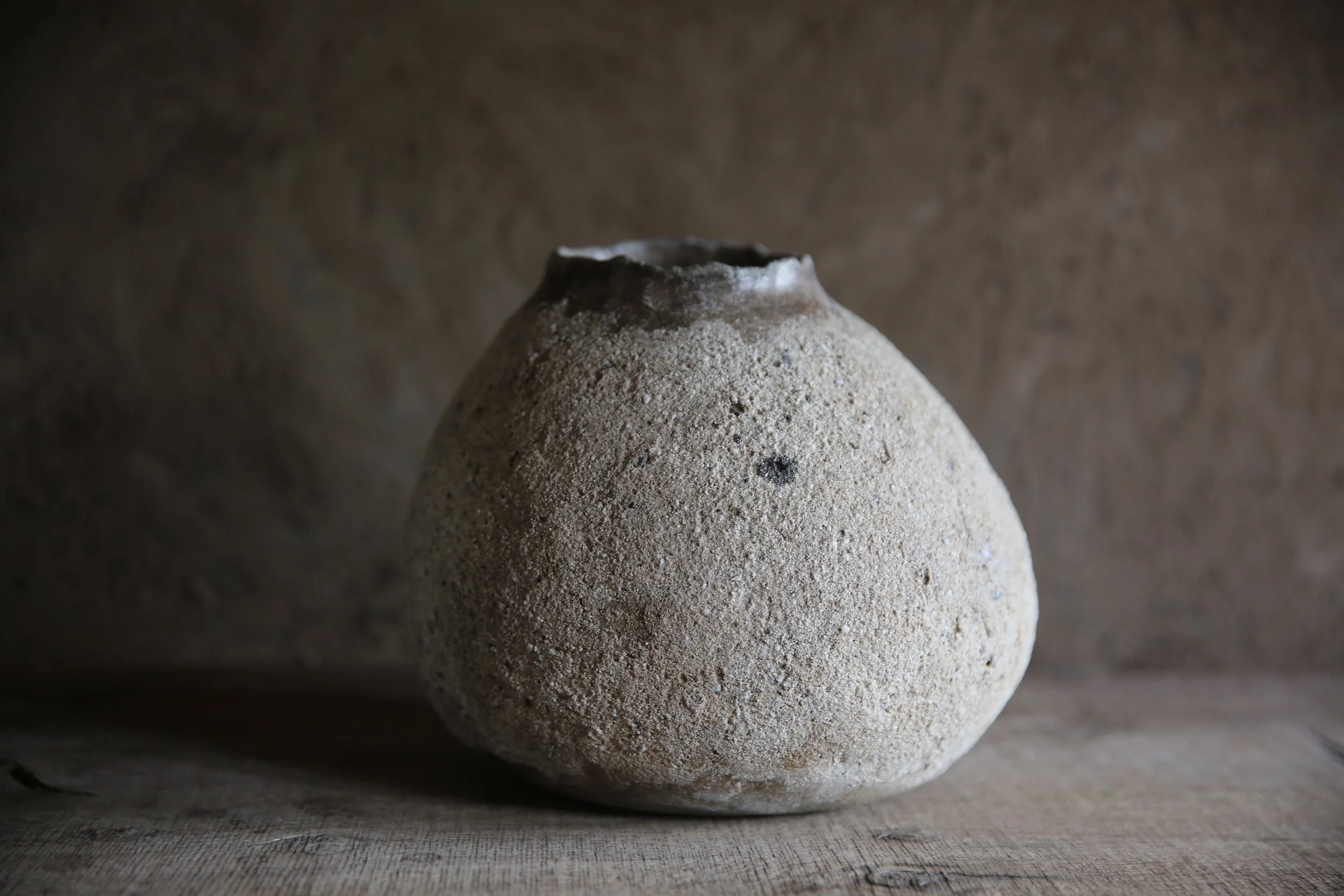 A textured ceramic vase with a rounded body and narrow opening, placed on a wooden surface against a brown background.