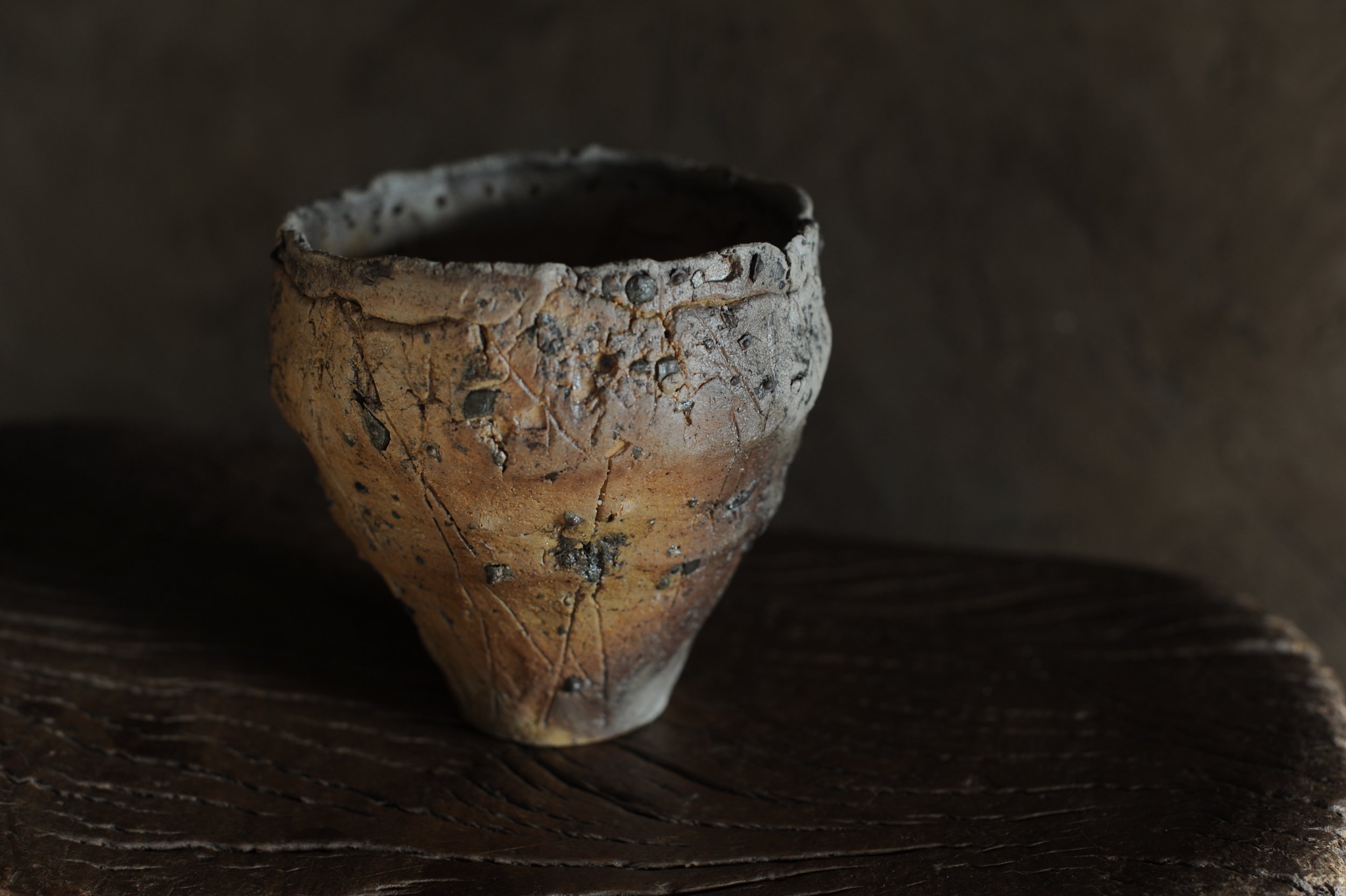 A small, rustic pottery cup with a textured surface and earthy tones, resting on a dark wooden surface against a dark background.