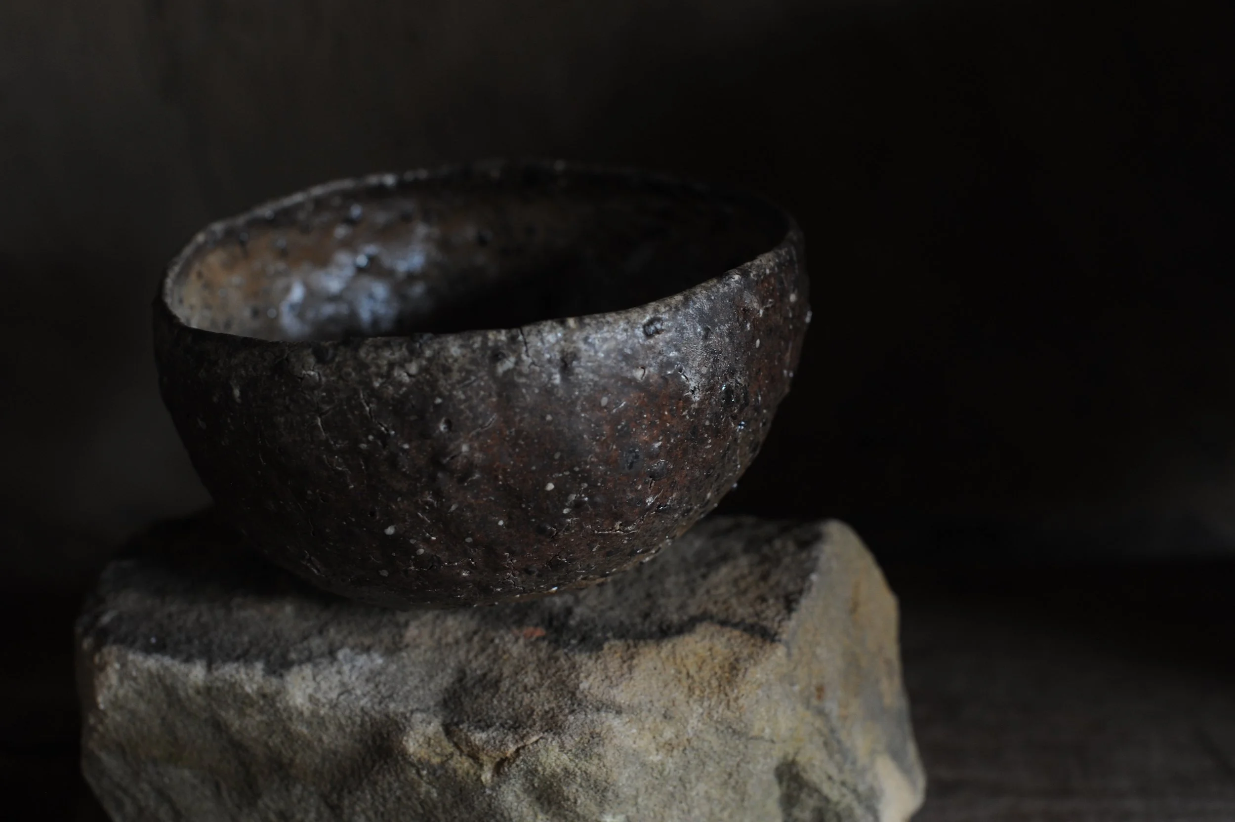 A rustic, dark brown ceramic bowl with an uneven, textured surface sitting on a rough stone against a black background.