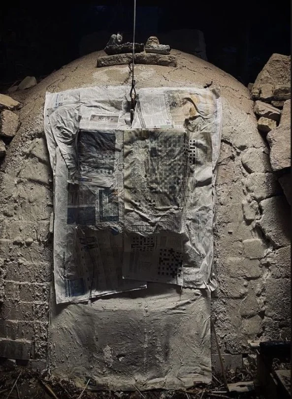 A brick and stone wall with a large, partially excavated, rounded stone structure at its center. Newspapers are taped on the structure, covering part of it, and there is a rope hanging vertically in the middle of the structure.