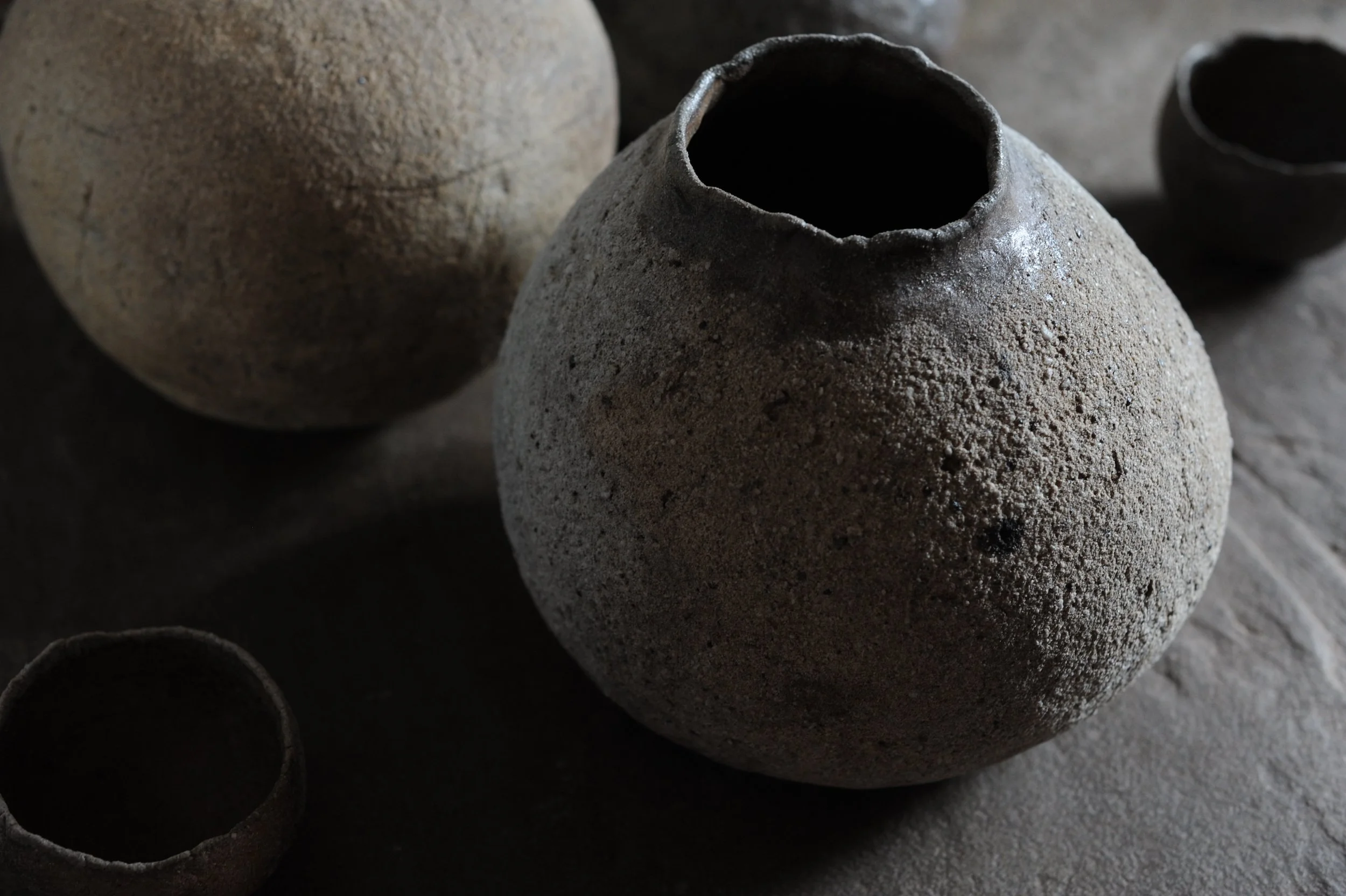 Several rustic, textured clay pots, with one large round vessel in the center and smaller pots surrounding it, placed on a wooden surface.