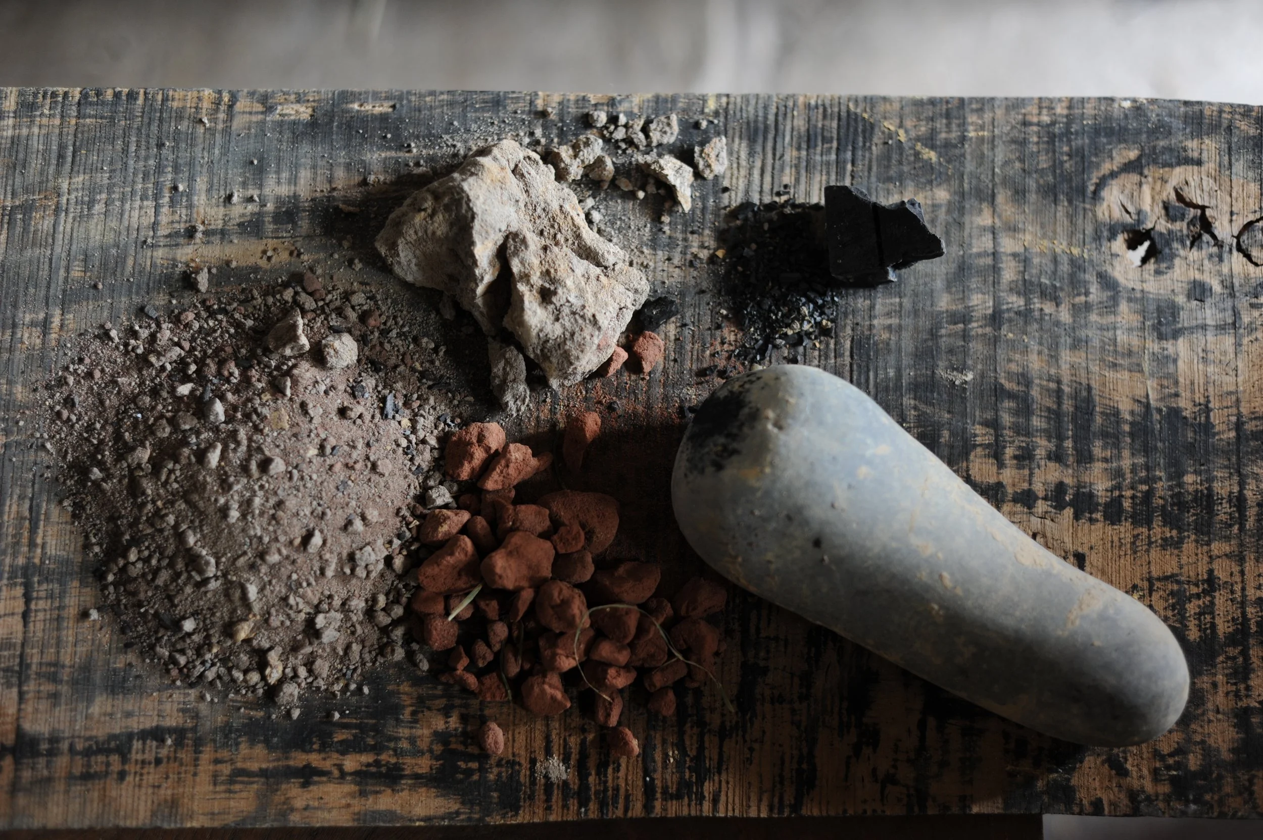 A variety of geological materials on a wooden surface, including rocks, gravel, charcoal, and a pestle.