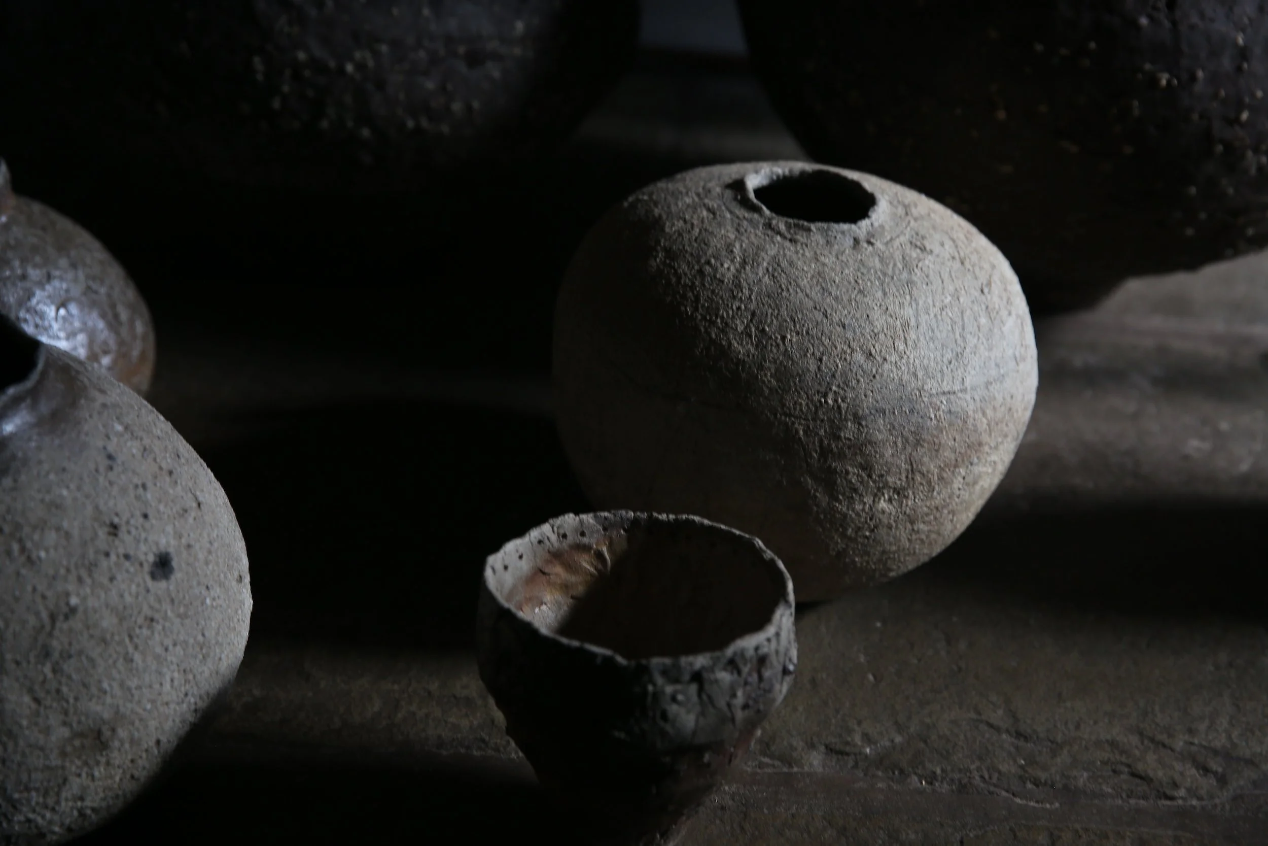 Several ancient pottery vessels, including a large round vessel with a hole at the top, and a smaller broken bowl, arranged on a dark surface with dramatic lighting highlighting their rough textures.