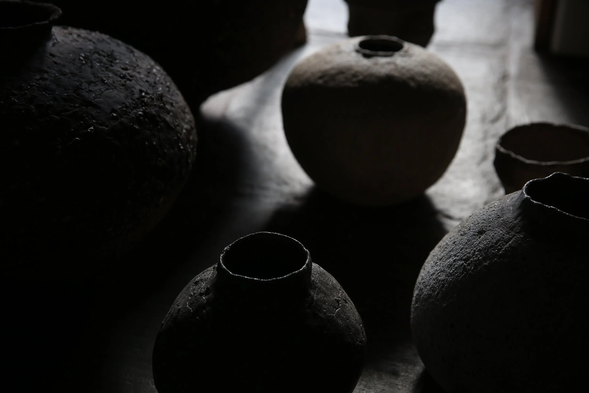 Dark, textured pottery jars and vases arranged on a surface, with some illuminated by light coming from the side.