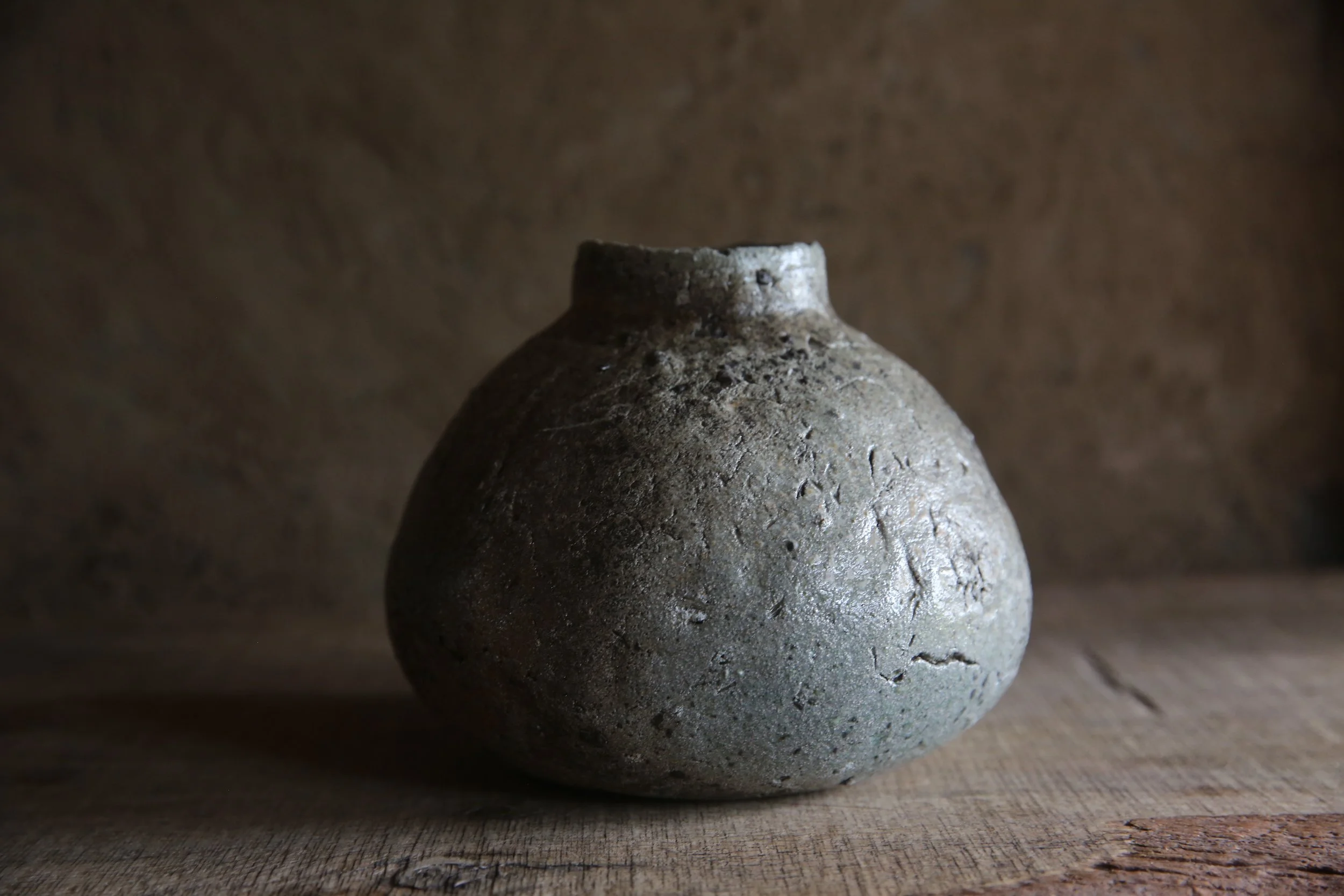 Ancient gray ceramic pot with rough texture on a wooden surface, with a plain brown background