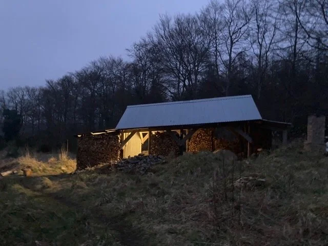A rustic barn with a metal roof, illuminated from inside, surrounded by trees and uneven grassy terrain at dusk.