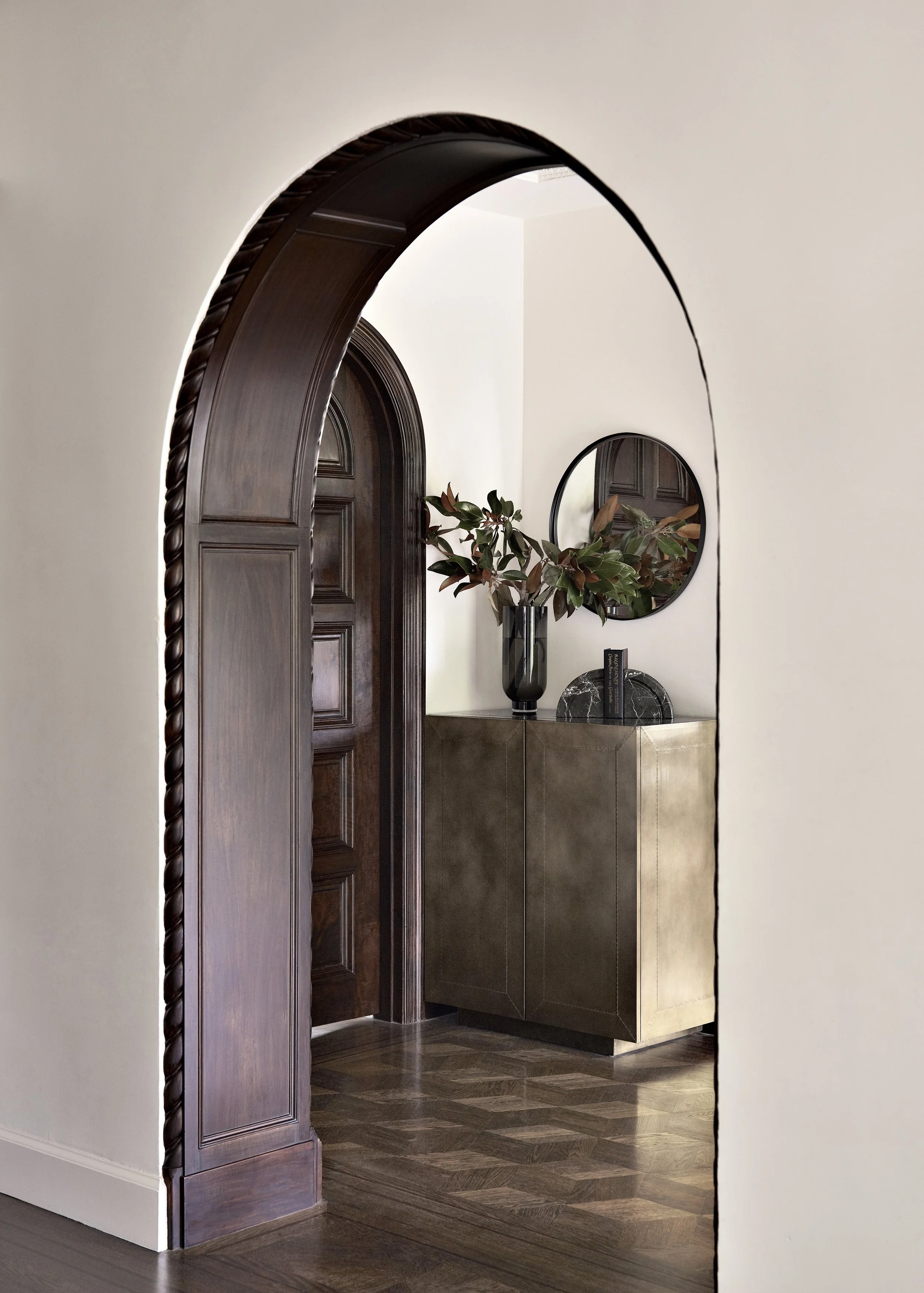 Living room with high, vaulted ceiling and wooden beams, featuring a chandelier, large window with curtains, a curved sofa with cushions, a piano, a round coffee table with a plant, and hardwood flooring with a rug.