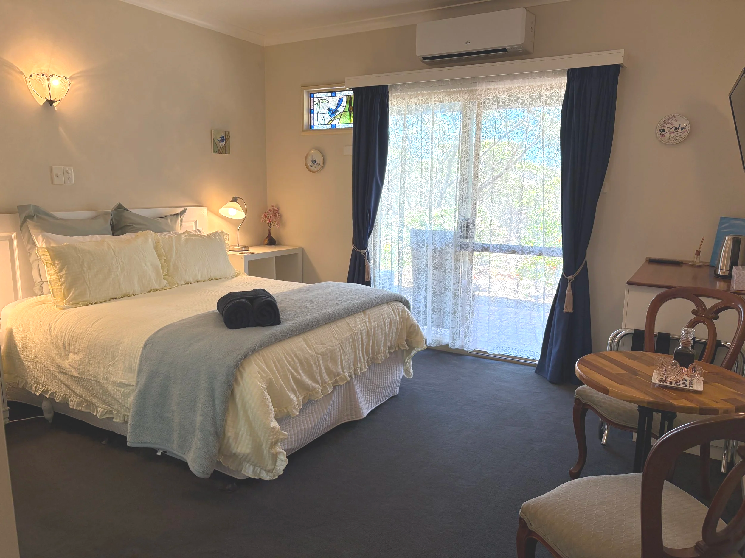 A cozy bedroom with a large bed covered in beige bedding and pillows, a nightstand with a lamp and pink flowers, a sliding glass door with lace curtains and blue drapes leading to a balcony, a small round wooden table with chairs, and a wall-mounted 