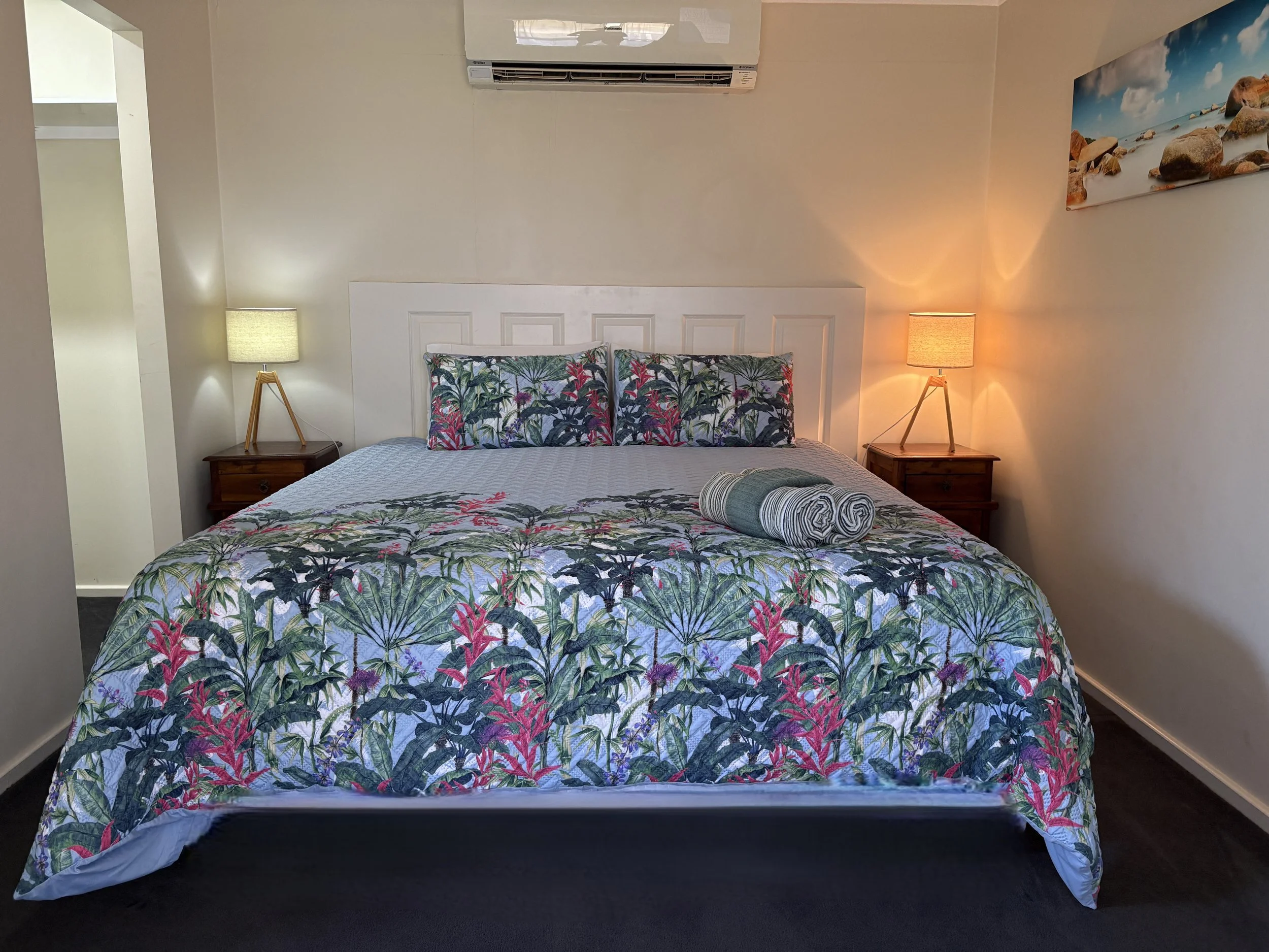 A neatly made bed with a tropical floral bedspread, matching pillows, and rolled towels on top, flanked by two wooden nightstands with lamps. There is a wall-mounted air conditioner above the bed and a seaside landscape photograph hung on the wall.