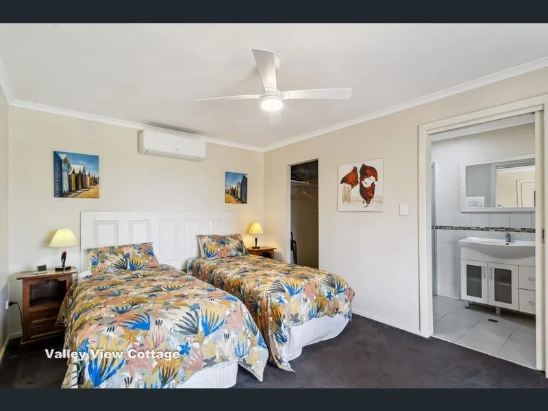 A bedroom with two queen beds covered in floral bedding, two nightstands with lamps, wall art, an air conditioner, and an open door leading to a bathroom with sink and mirror.