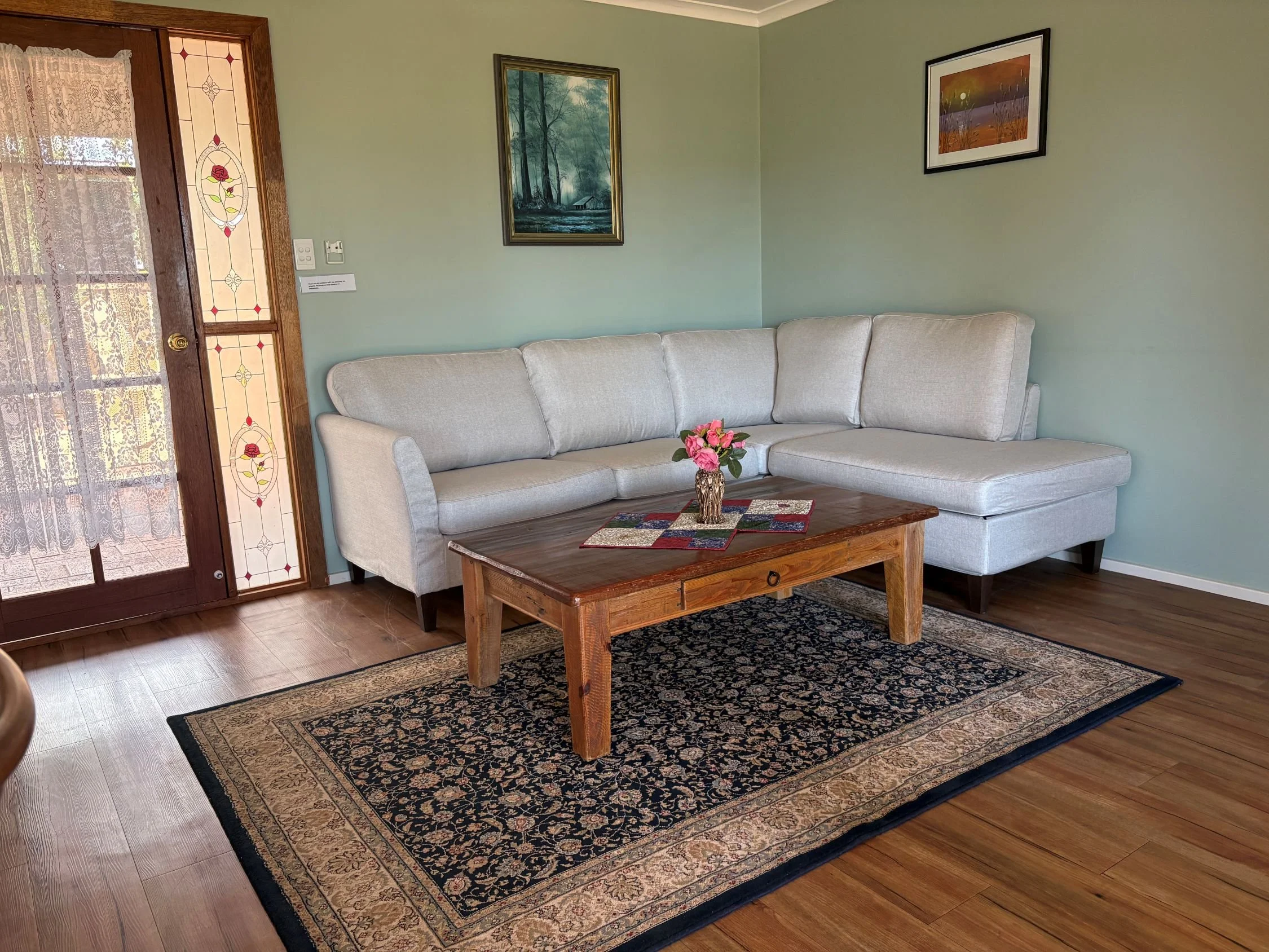 Living room with a beige sectional sofa, a wooden coffee table with a pink flower arrangement, a patterned area rug, artwork on light green walls, and a door with stained glass and lace curtains.