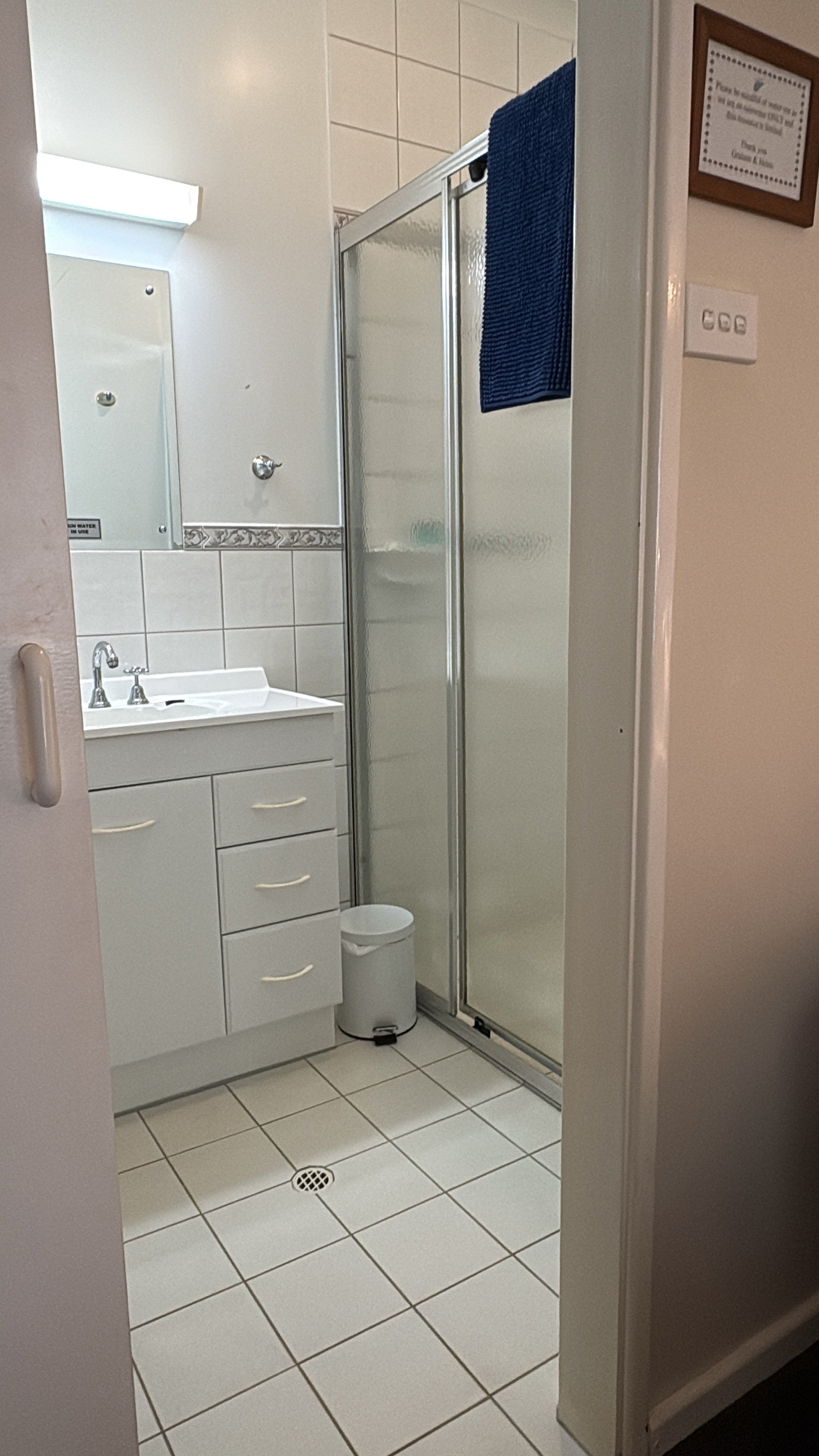 A small bathroom with a white vanity sink, a mirror with a light above it, a shower with a frosted door, a blue towel hanging on the shower door, a small white trash can, and a framed certificate or sign on the wall.