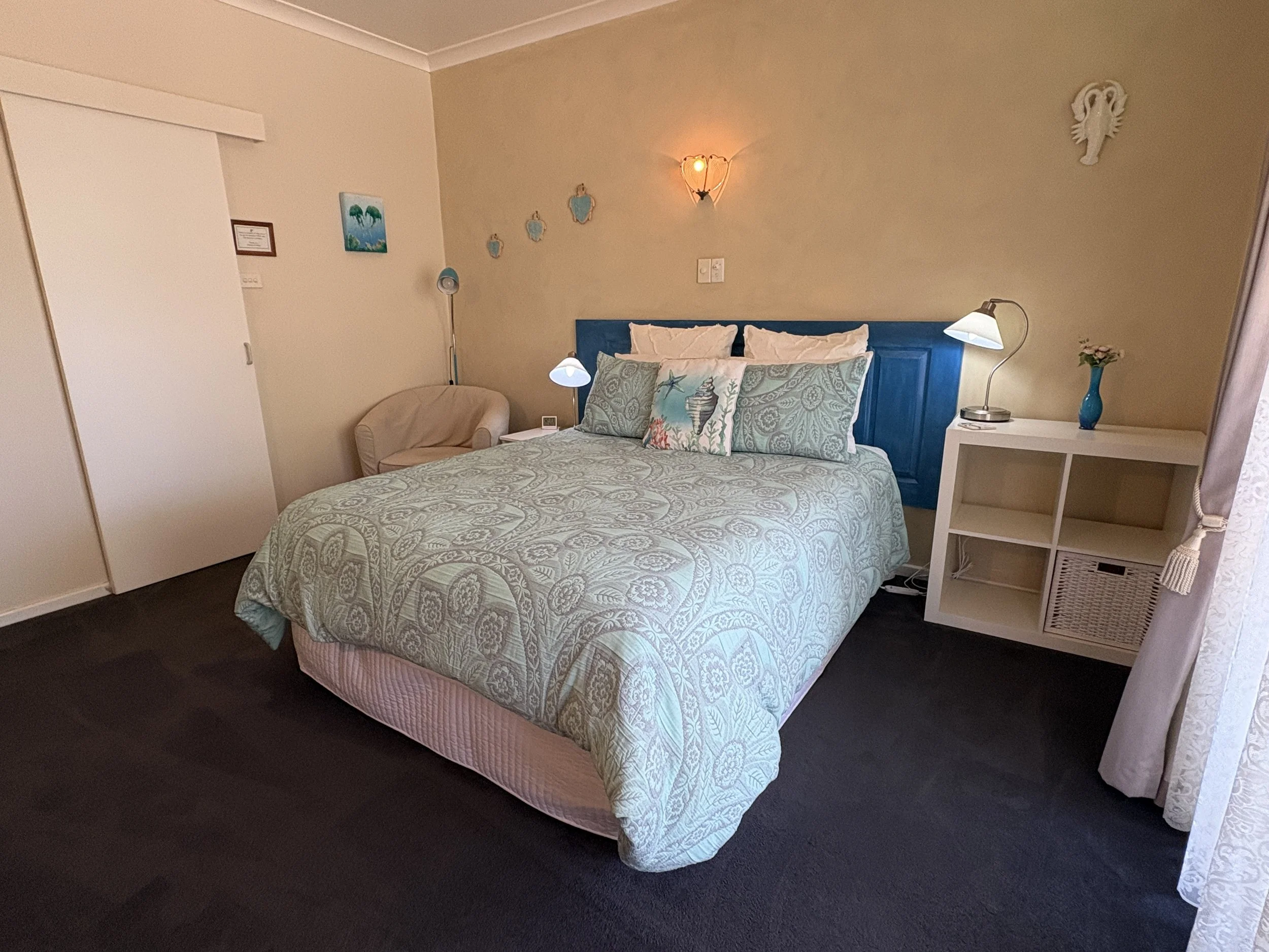 A cozy bedroom with a bed featuring a blue headboard, light blue and white bedding, and assorted pillows. There is a white side table with a lamp and a blue vase with flowers, and a white shelving unit. A beige armchair sits beside a floor lamp, and 