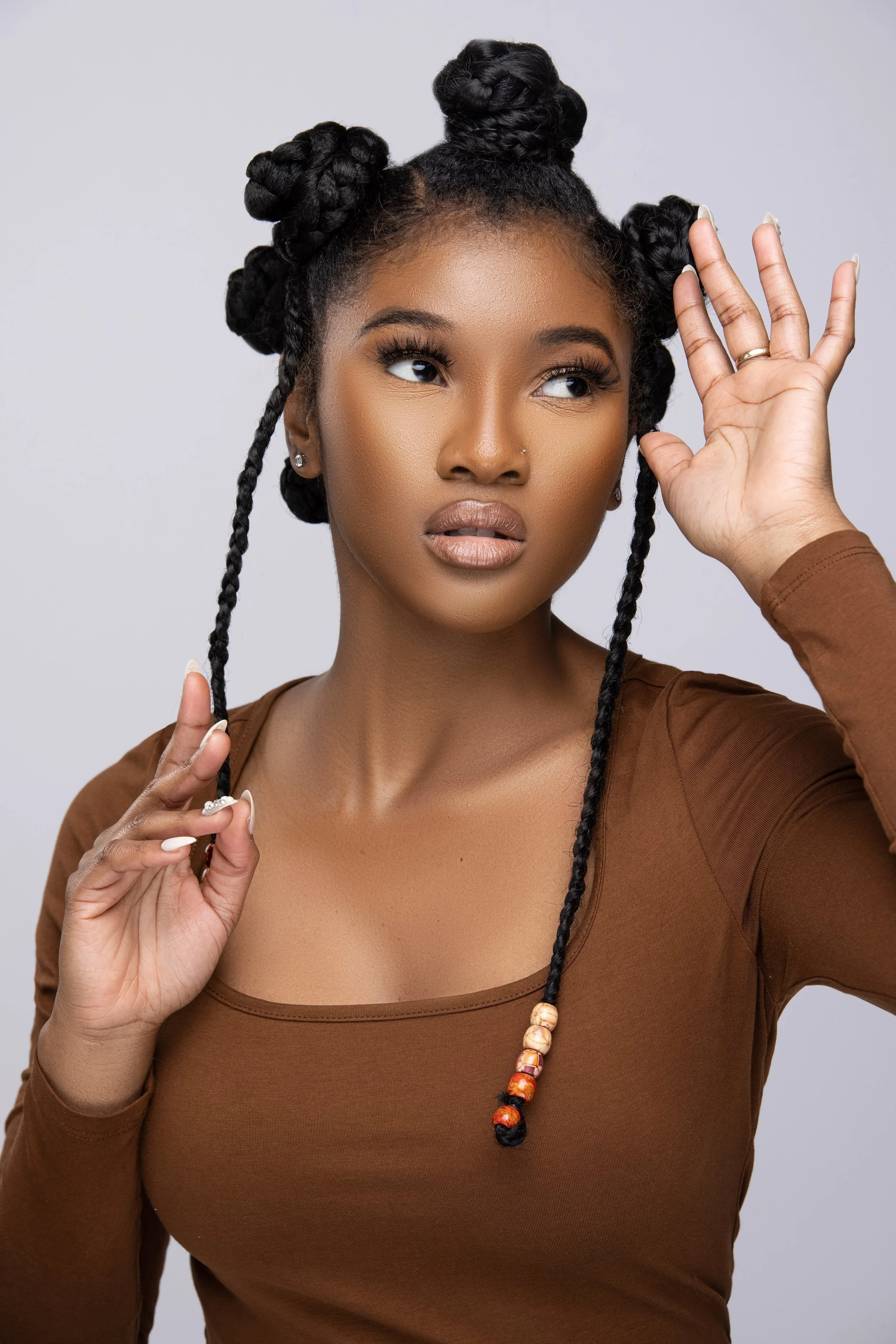 A woman with styled hair in Bantu knots with beads, wearing a brown top, holding a lock of her hair, and making a gesture near her face.