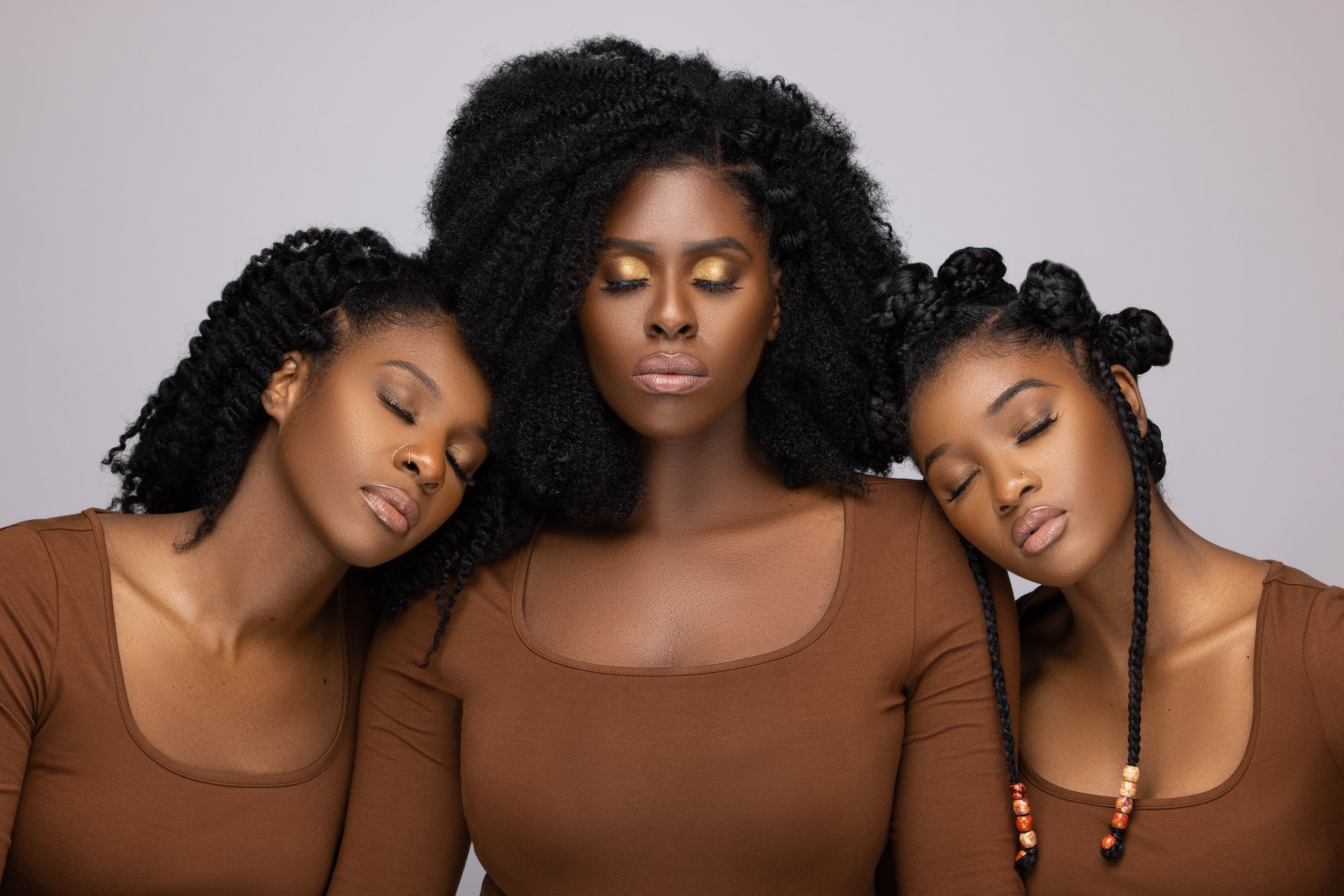 Three women with curly hair and brown tops, with their eyes closed, standing close together against a gray background.