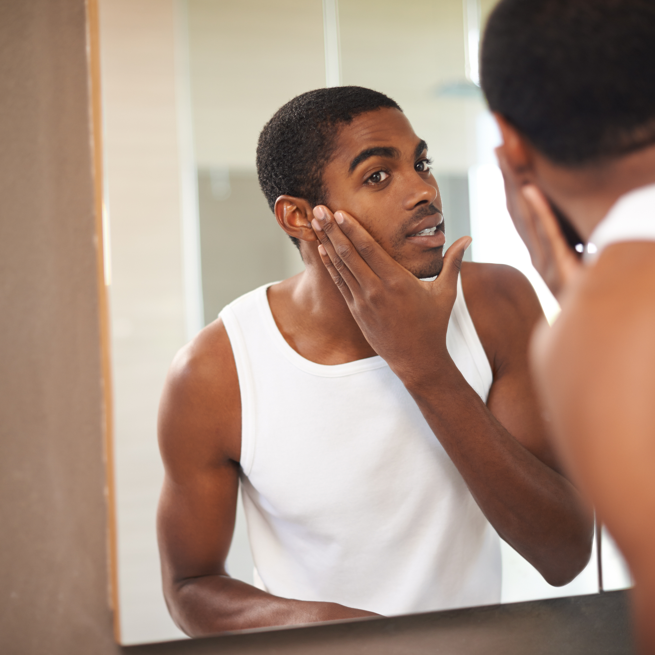 A young man wearing a white tank top looking at himself in the mirror while touching his face.