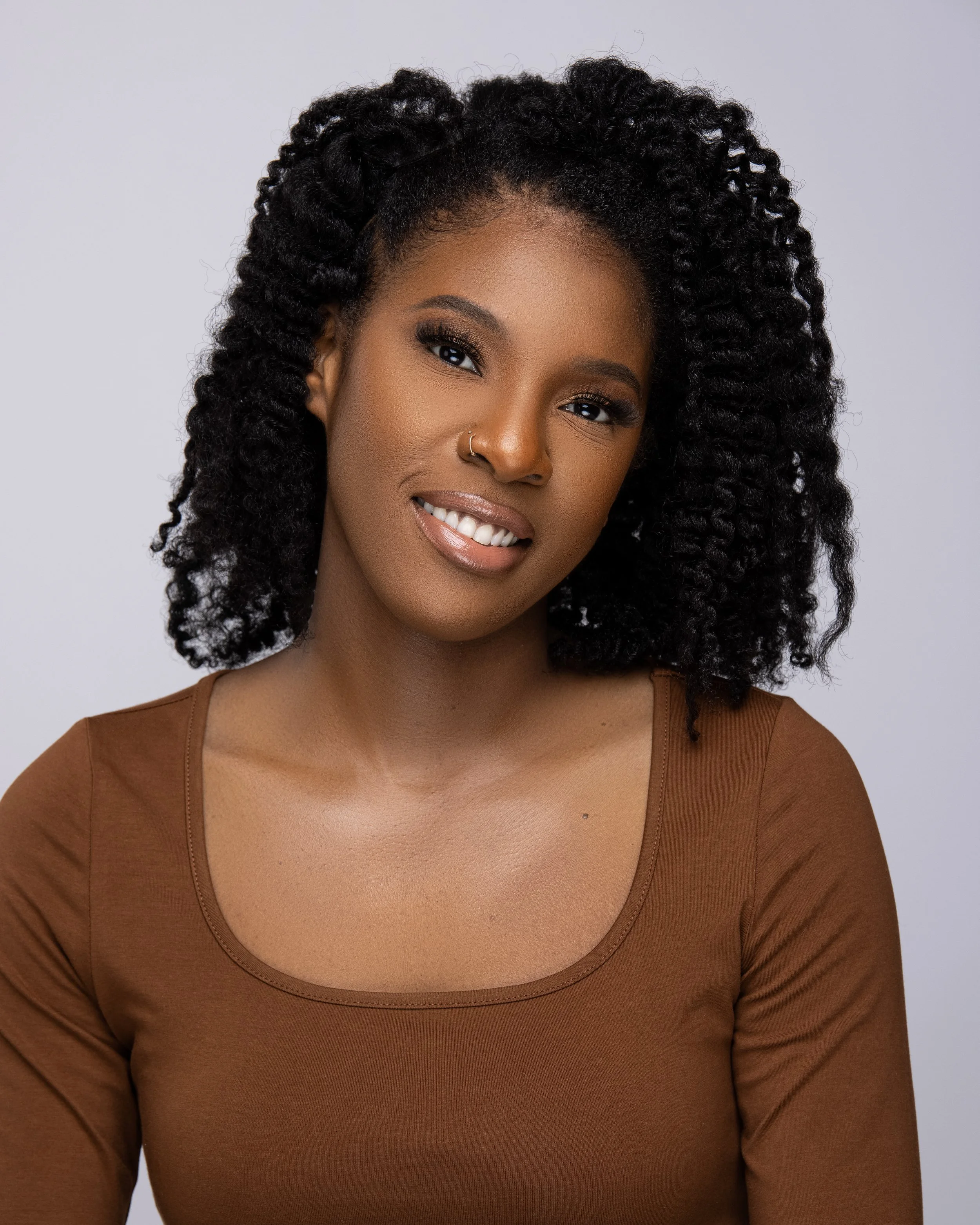 Portrait of a woman with curly hair, wearing a brown top, and sporting a nose ring, smiling slightly against a plain light gray background.