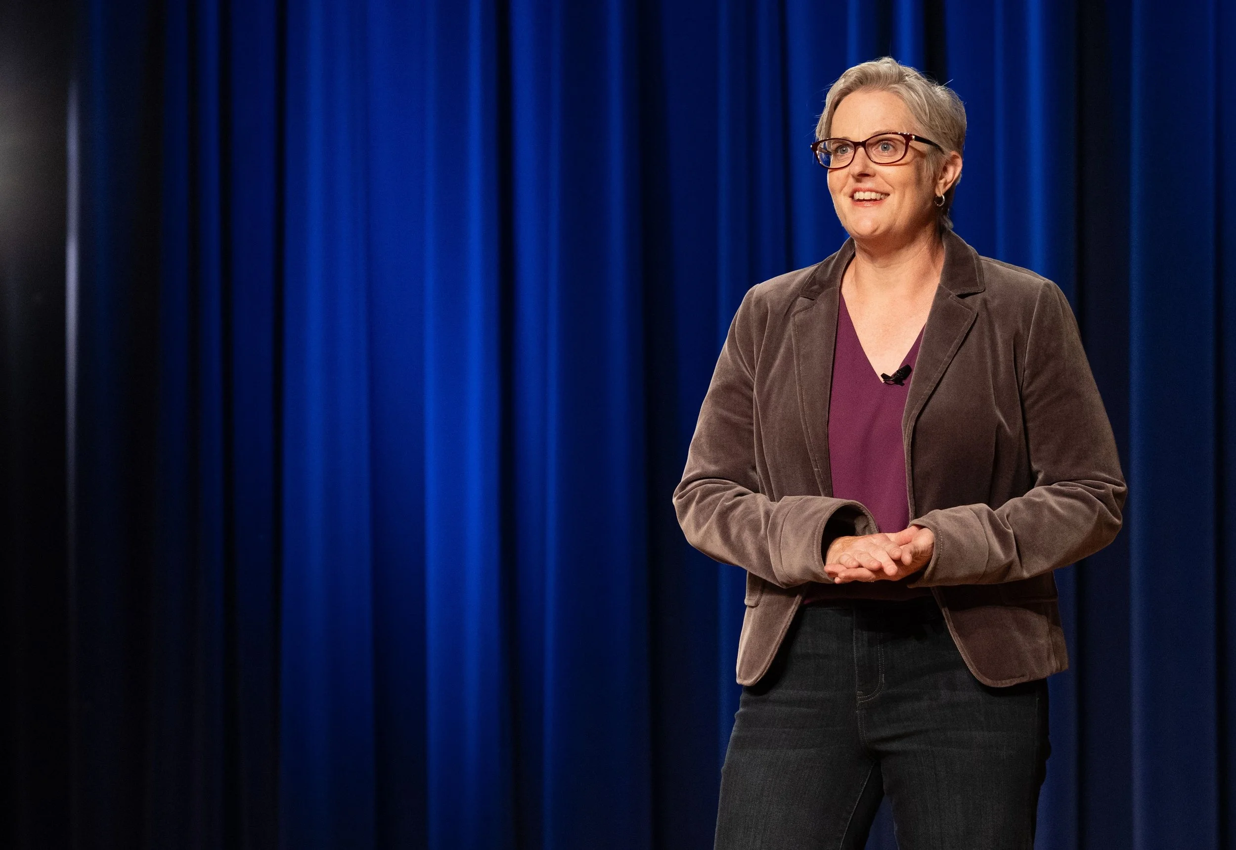 bernadette smith, a white woman with short blonde hair and glasses smiles on stage in a blazer and jeans.