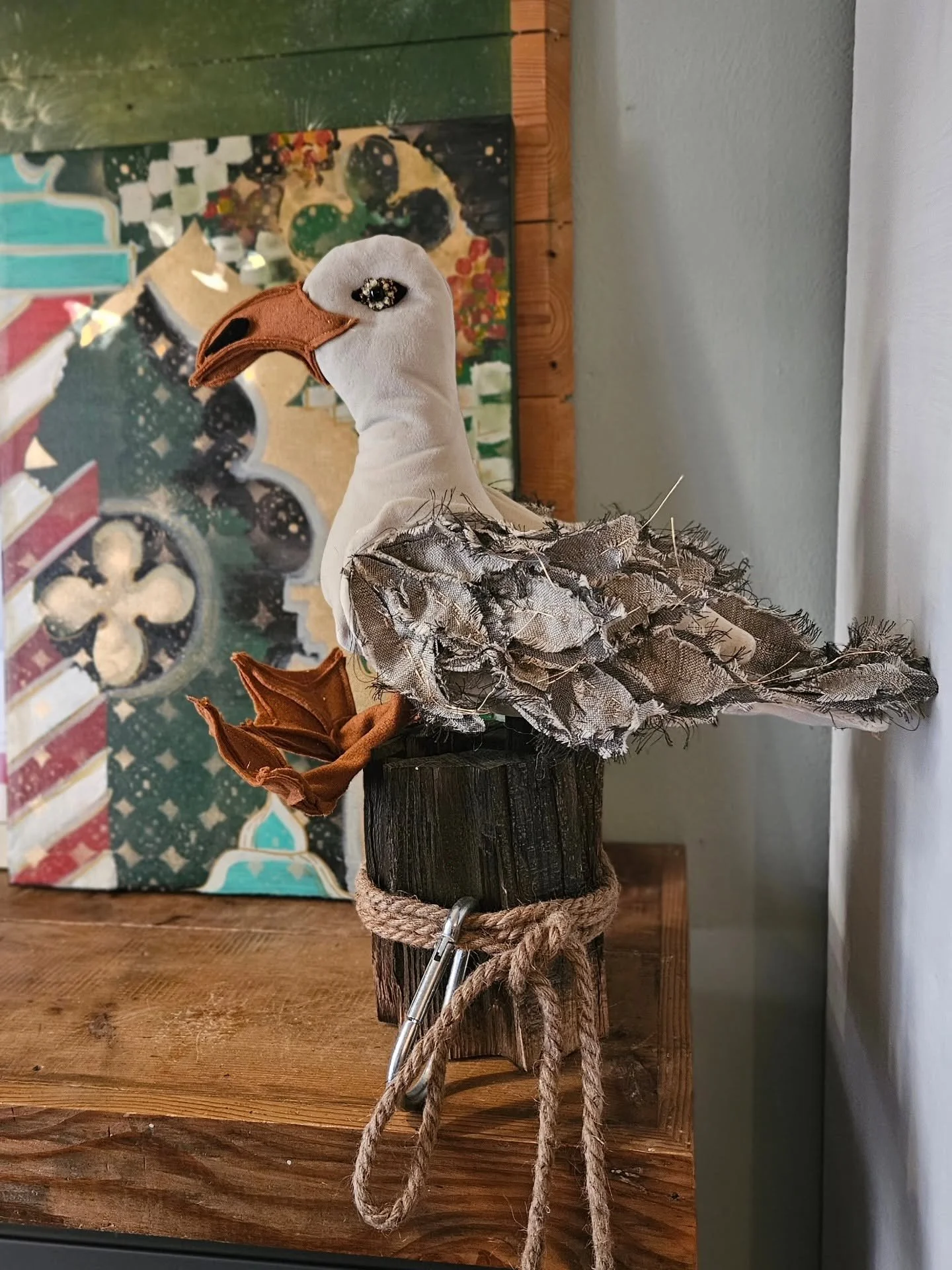 Una scultura di un uccello, probabilmente uno storno, realizzata con stoffe e materiali vari, posata su un tronco di legno e decorata con una corda di iuta. Sullo sfondo, un quadro colorato e il muro bianco.