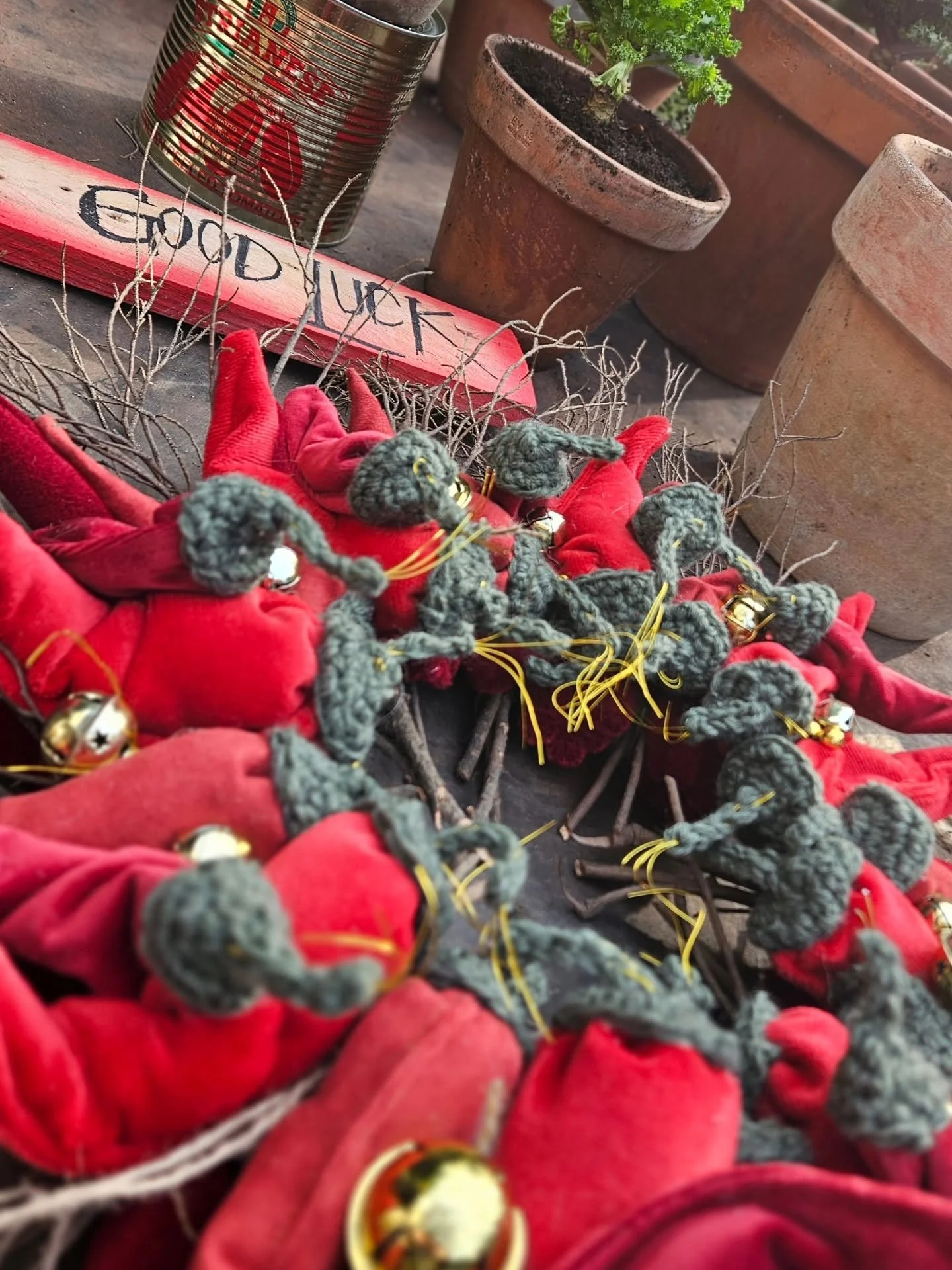 Bonfire setup with red fabric wreaths decorated with small golden bells, surrounded by terracotta plant pots, a can, and a painted wooden sign that says 'Good Luck' in English.