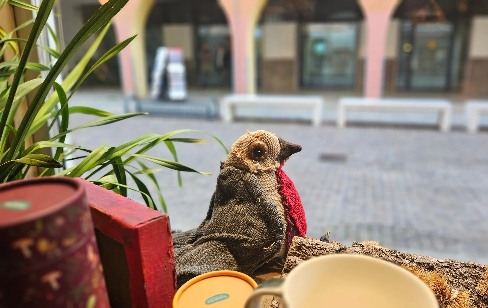 Una marionetta di un uccello con tessuto marrone, nero e rosso, posizionata tra piante e oggetti in primo piano, con uno sfondo di negozio con vetrine e edifici.