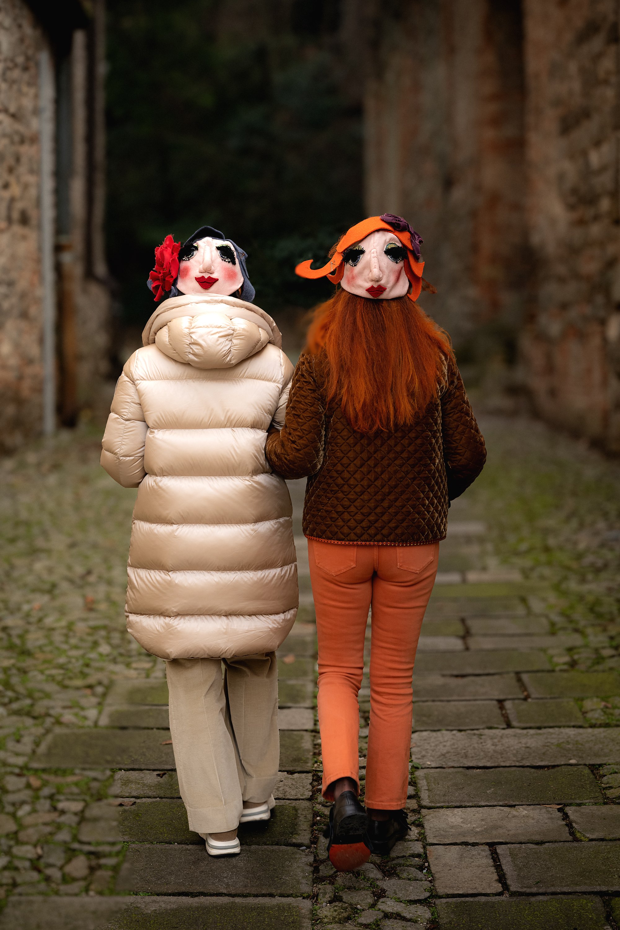 Due persone con maschere di marionette che camminano insieme in un cortile con pavimentazione in pietra, circondato da muri di mattoni.