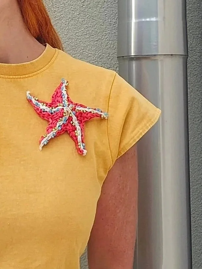 Particolare di una persona con una maglietta gialla e una spilla a forma di stella marina decorata con stoffe colorate e ricami.