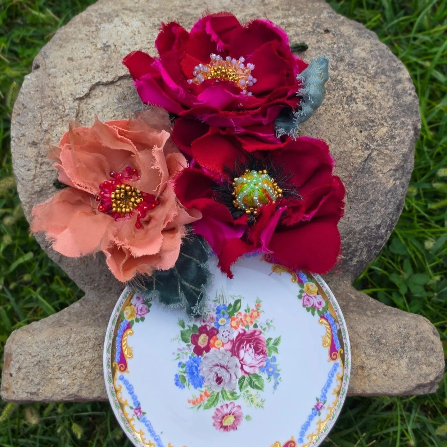 Fiori artigianali di stoffa di vari colori, decorati con perline, posati su una pietra, con una piattina con fiori dipinti al centro.