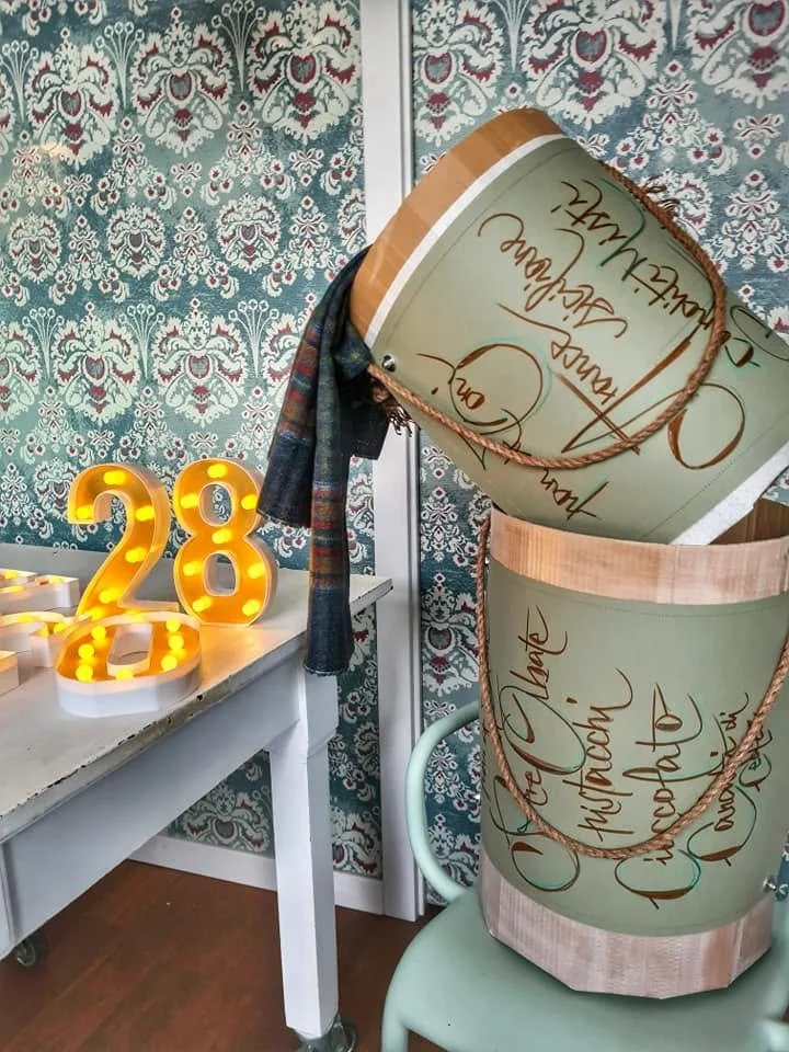 Decorazione di compleanno con le candeline luminose a forma del numero 28, un grande cartone decorato con scritte in italiano, un panno a scacchi appoggiato su una sedia, e un tavolo bianco con dettagli ornamentali sullo sfondo.