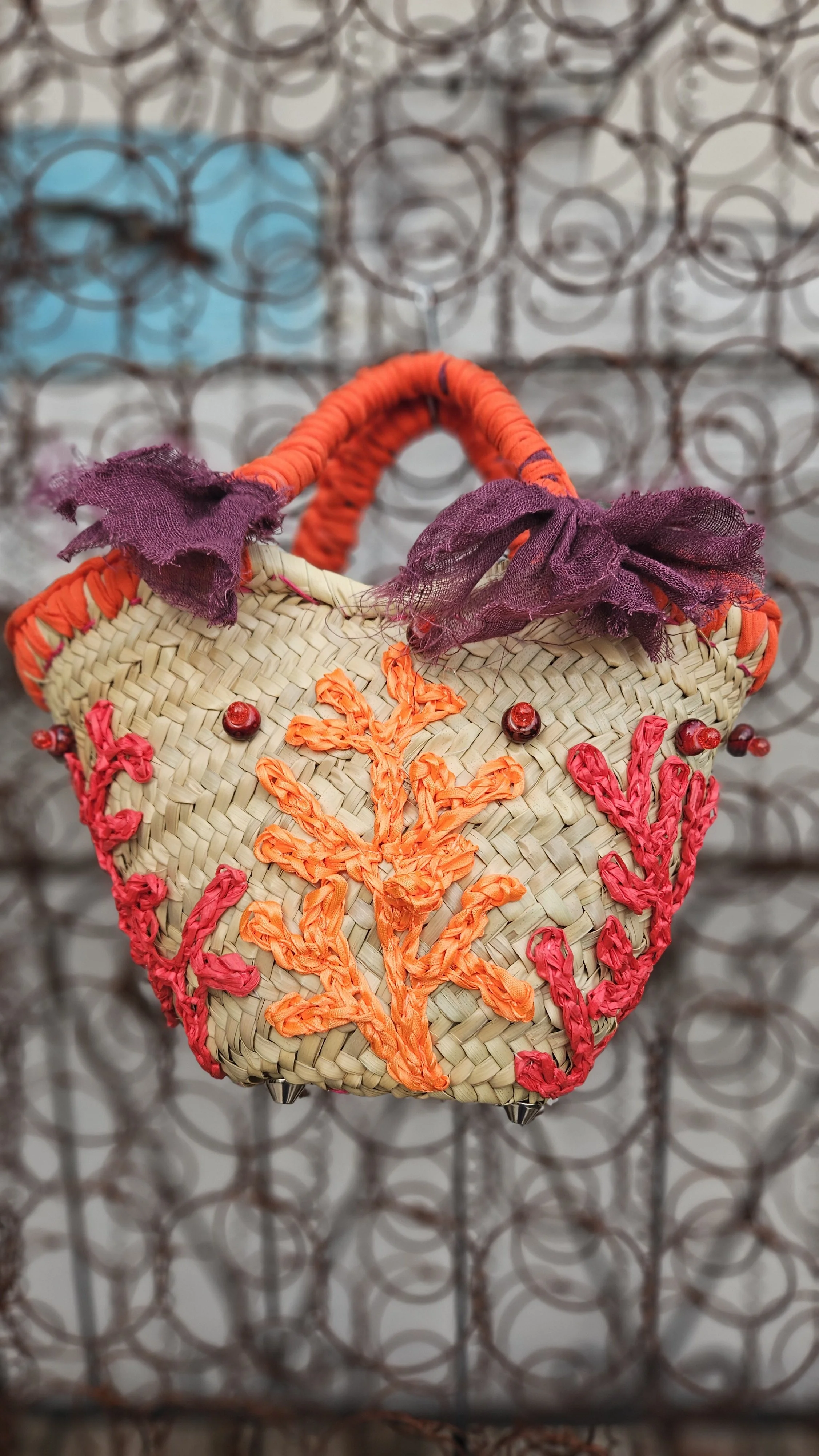Borsa decorata con fili di tessuto arancioni e rosa cuciti su una superficie di paglia, con dettagli di stoffa viola attorno alla maniglia, appesa a una rete metallica.