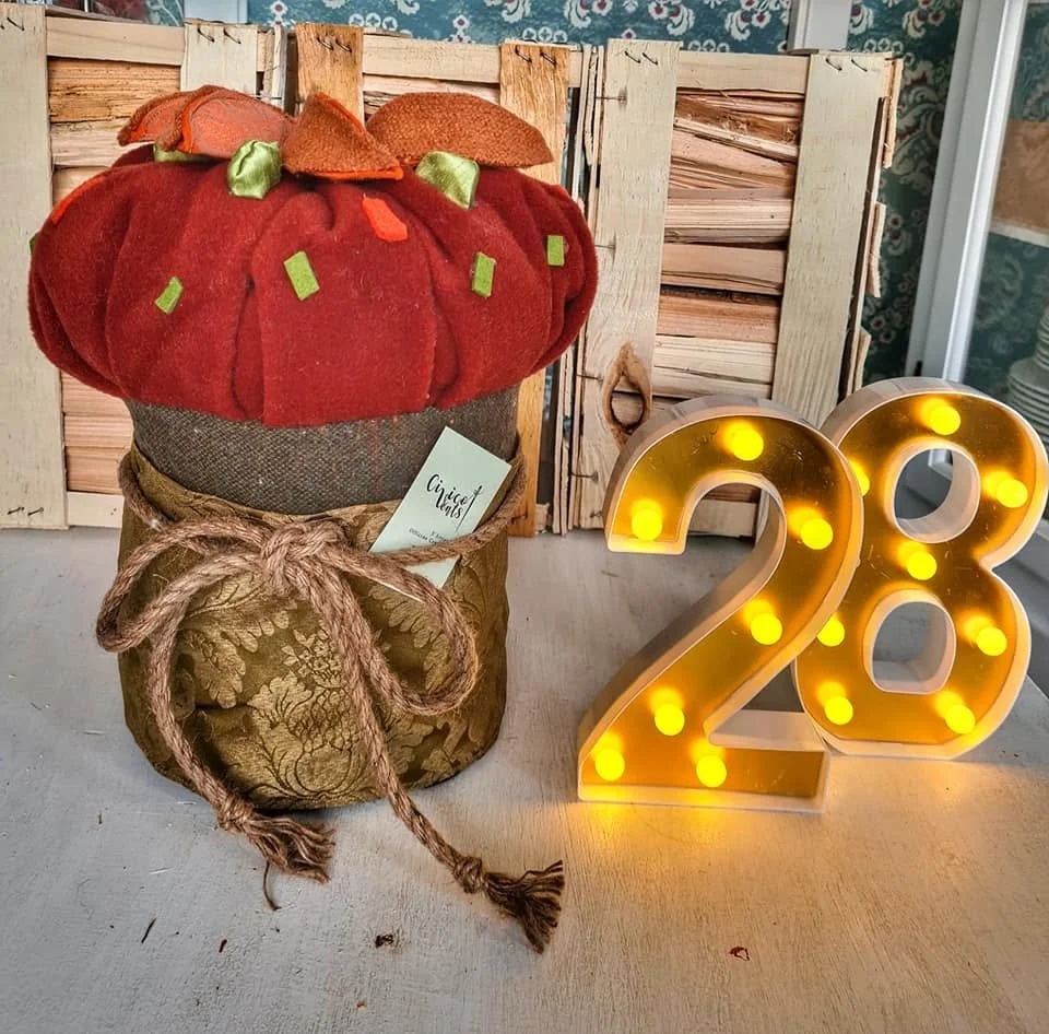 Festa di compleanno con decorazioni numeriche luminose '28' e un vasetto decorato con tessuto e corda, che somiglia a una zucca di stoffa con un feltro rosso sulla parte superiore, su sfondo di casse di legno e pareti con carta da parati blue.