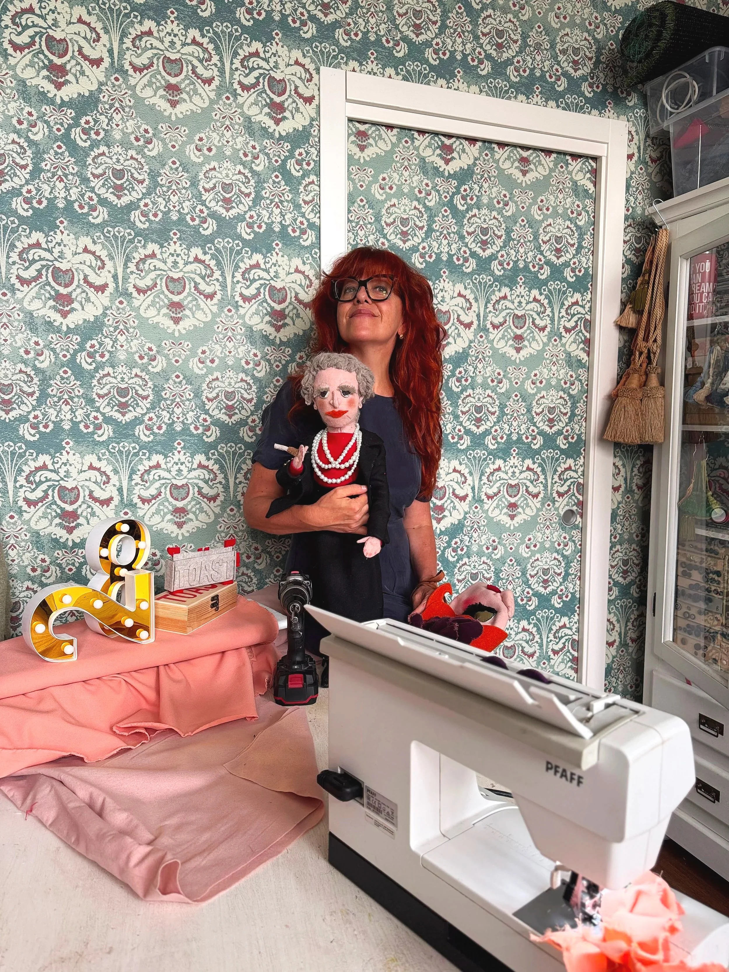 Donna con capelli rossi, occhiali e sorriso, che tiene una bambola con capelli grigi, denti rossi e collana di perle, in un ambiente decorato con carta da parati ornamentale. Davanti c'è una macchina da cucire bianca. C'è anche una poltrona rosa, decorata con un'illuminazione a lettere e vari oggetti, tra cui un telefono. Sulla destra, un armadietto con oggetti e tessuti.