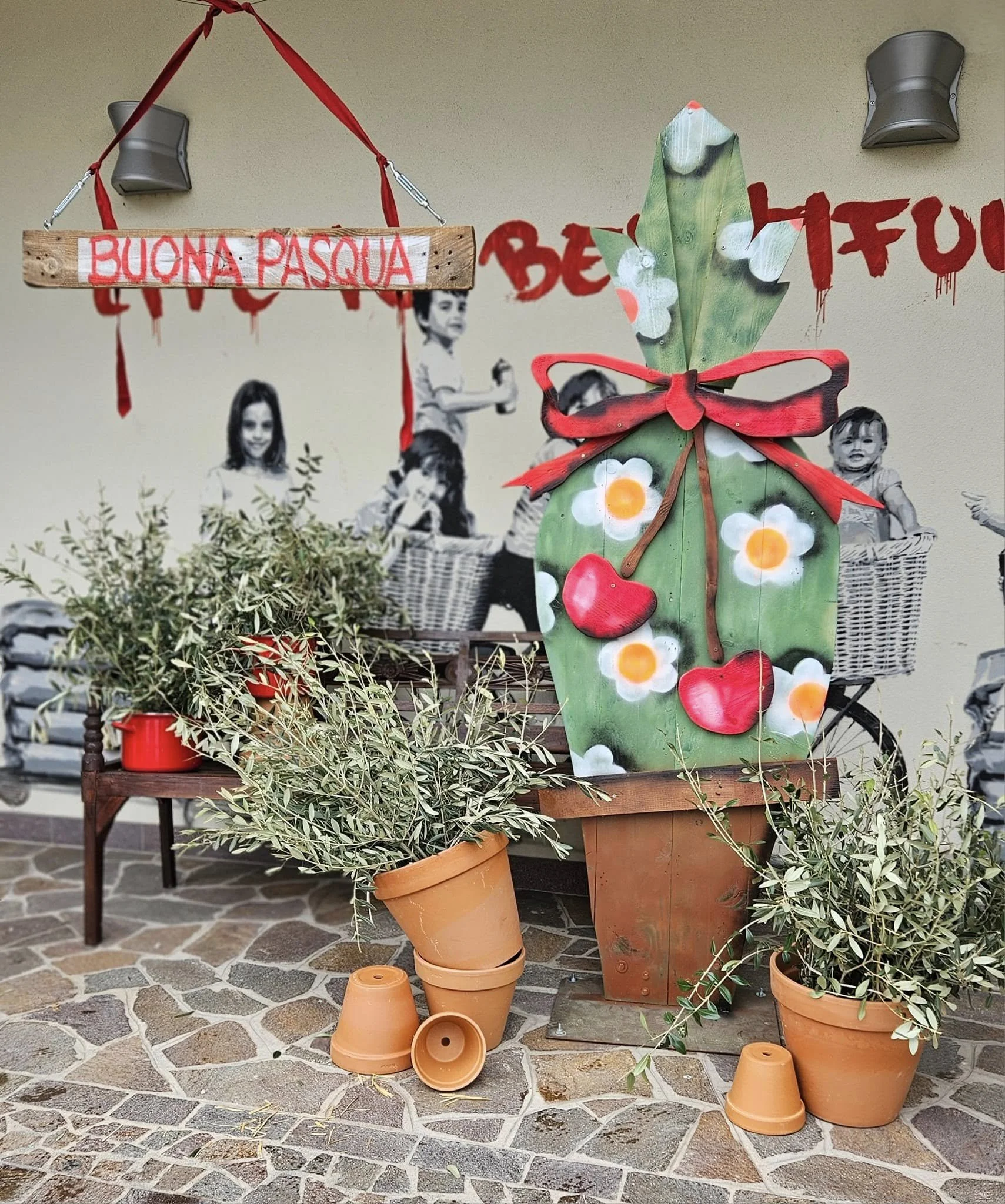 Decorazione pasquale con vasi di terracotta e piante, uova di carta colorate, scritta 'Buona Pasqua', e sfondo con disegni di bambini e decorazioni pasquali.