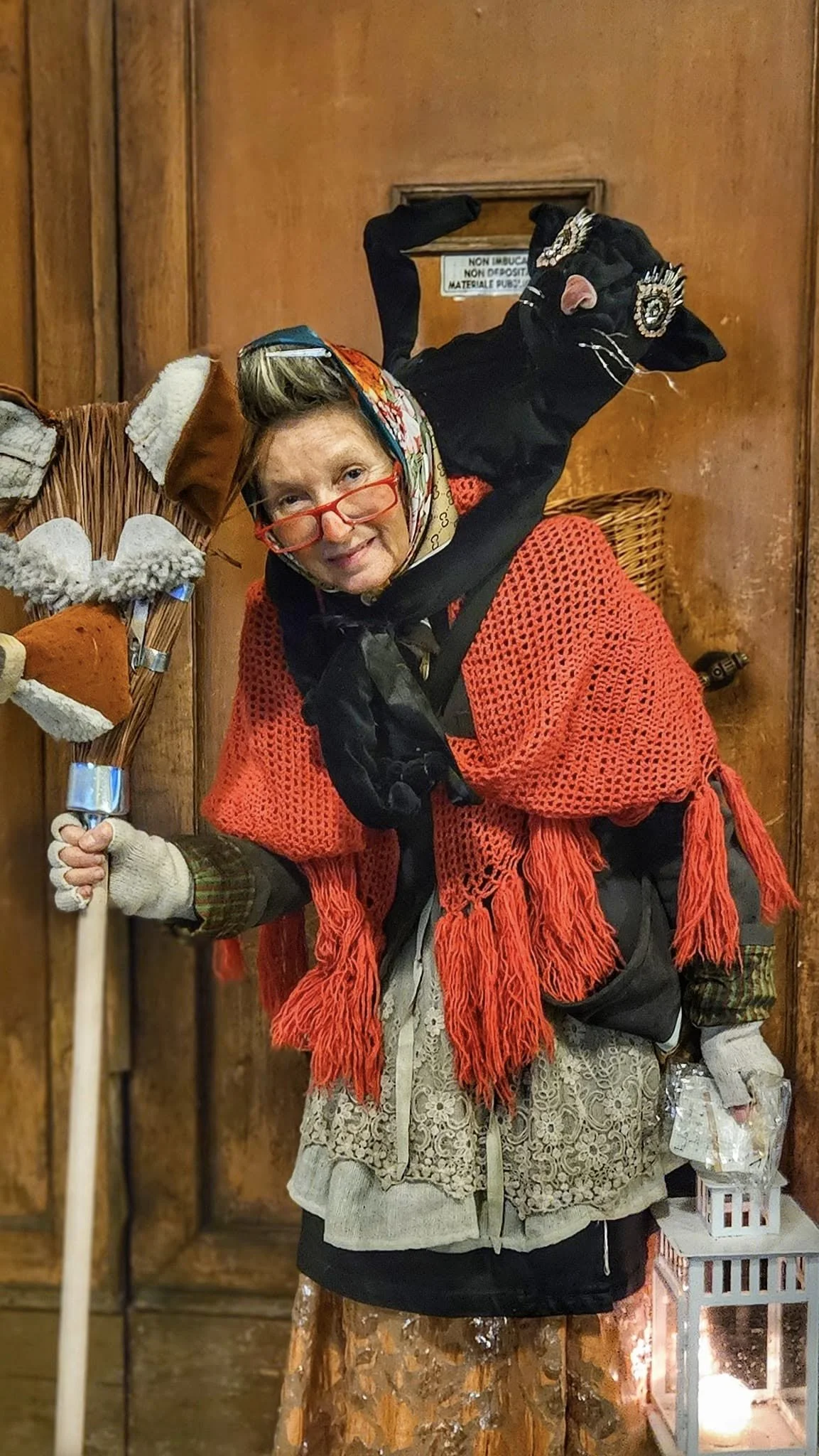 Una donna vestita con abiti e accessori vintage, con un cappello di pizzo e un foulard in testa, che tiene una scopa e un foglio in mano, e indossa occhiali rossi. Sullo sfondo un elemento di legno e due gatti neri, uno di fronte all'altra.