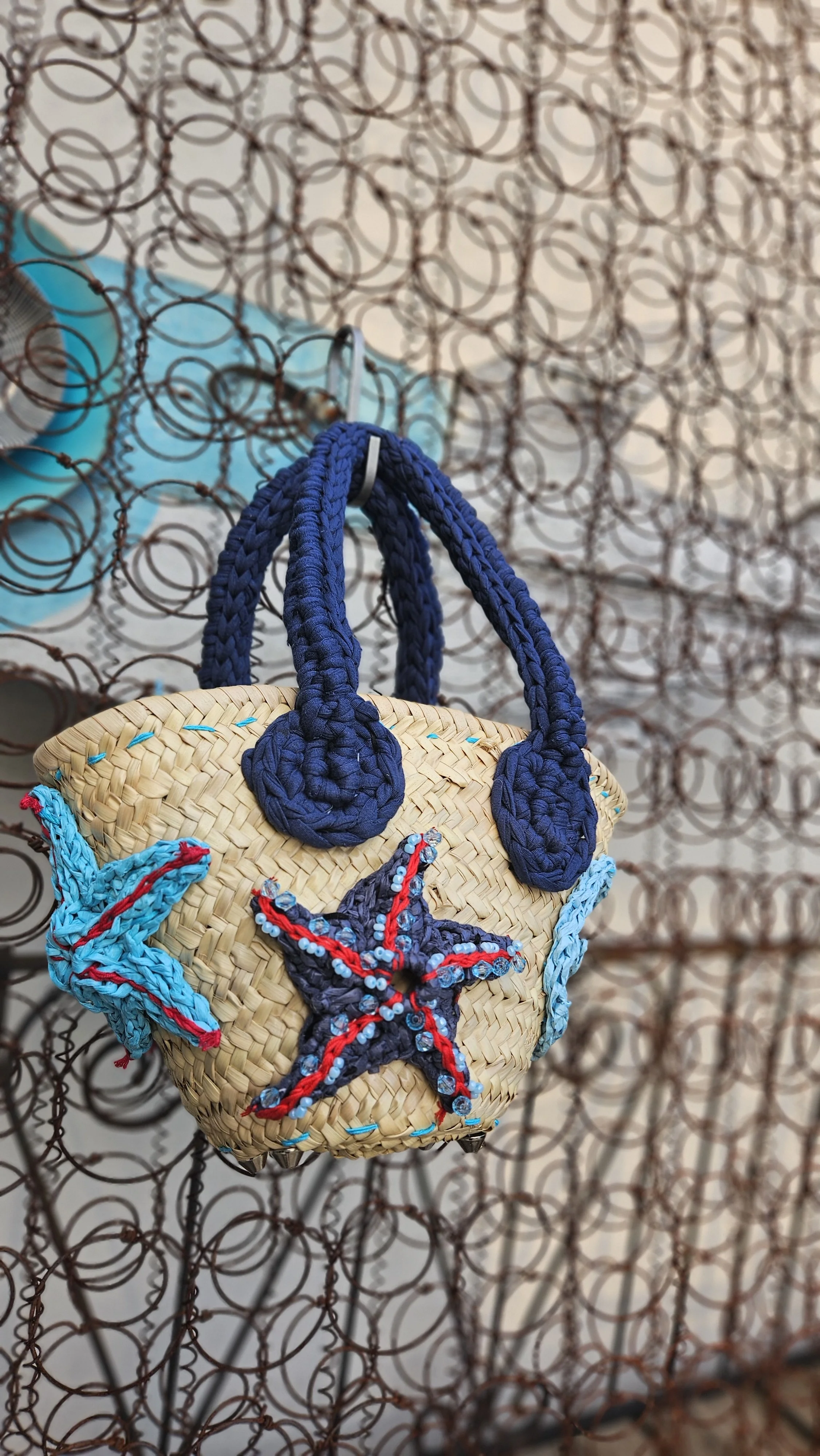 Una borsa di vimini decorata con ricami di stelle marina, appesa a una recinzione di ferro arrugginita.