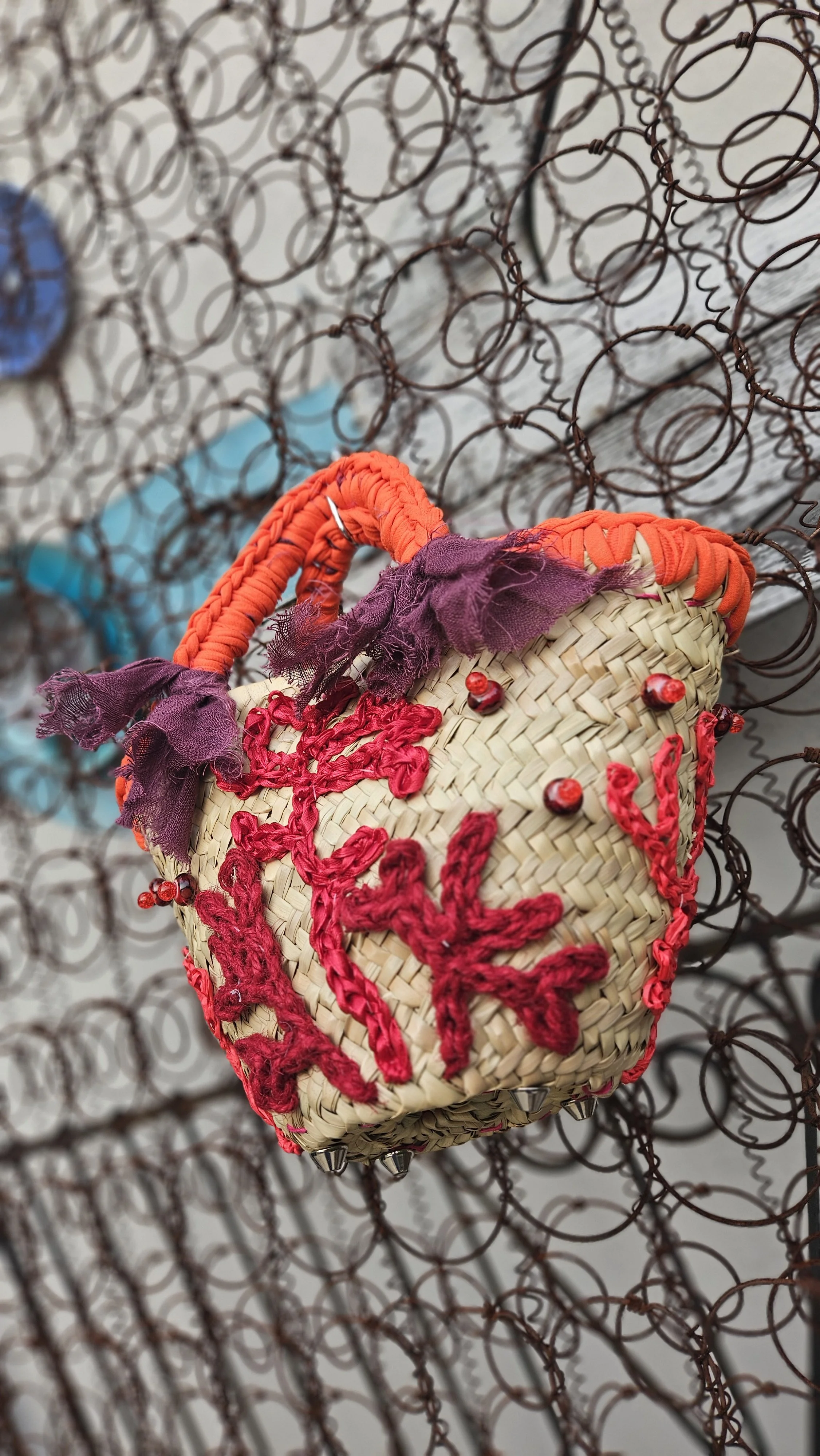 Borsa di paglia decorata con ricami rossi, fiori e foglie di tessuto viola, appesa a una rete di filo di ferro intrecciato.