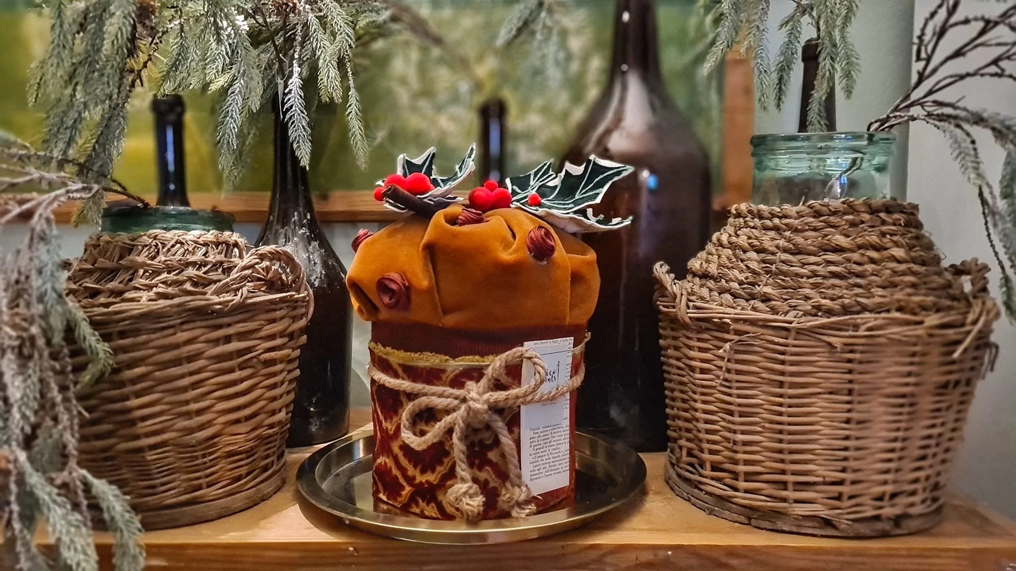 Decorazioni natalizie con vasi di vimini, bottiglie di vetro e un pacchetto regalo avvolto in stoffa arancione con fiocco di spago naturale, adornato con bacche rosse e foglie di agrifoglio.
